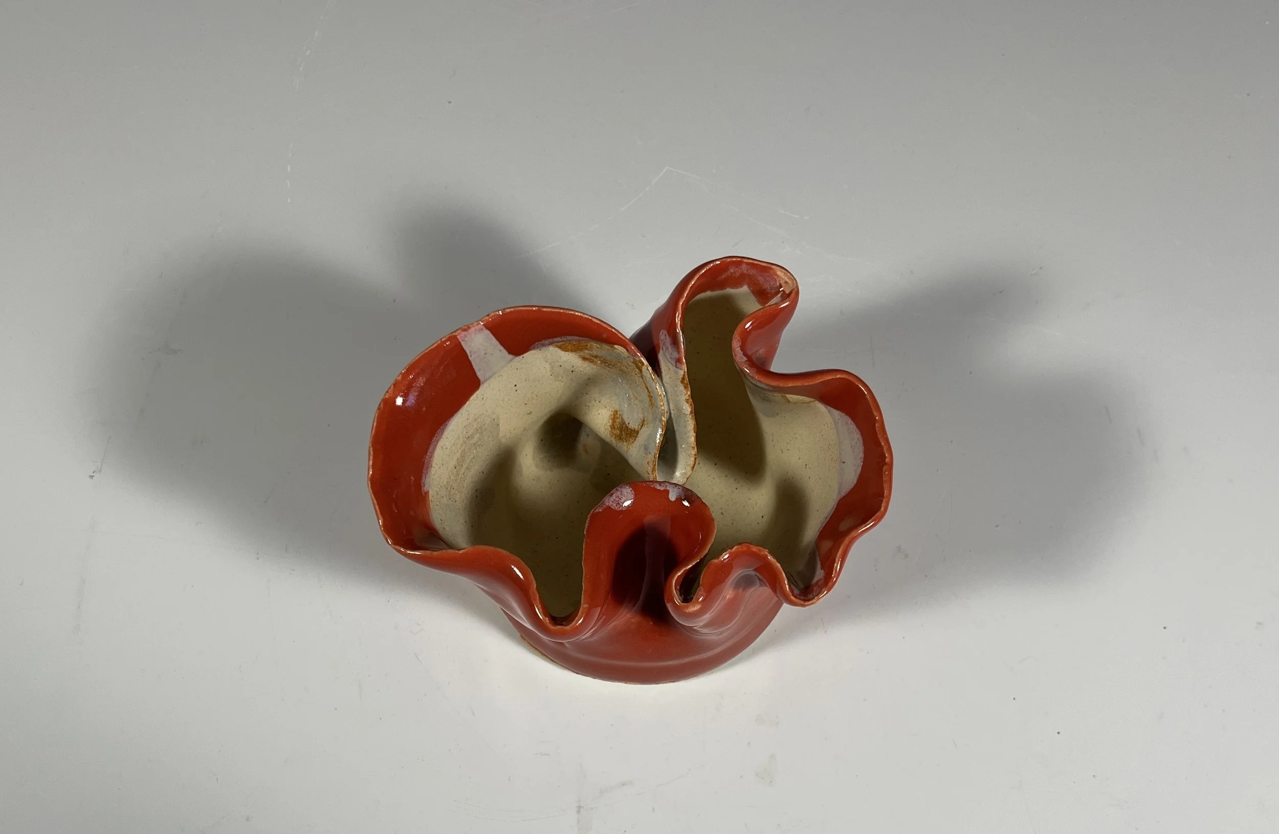 Ceramic bowl with a red, ruffled edge, beige interior, and irregular shape, casting a shadow on a white surface.