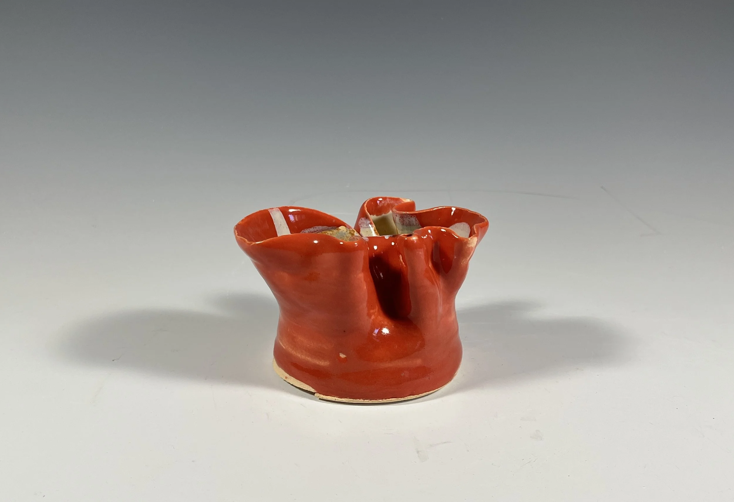 Small ceramic vase with glossy red glaze and irregular shape, placed on a plain white surface.