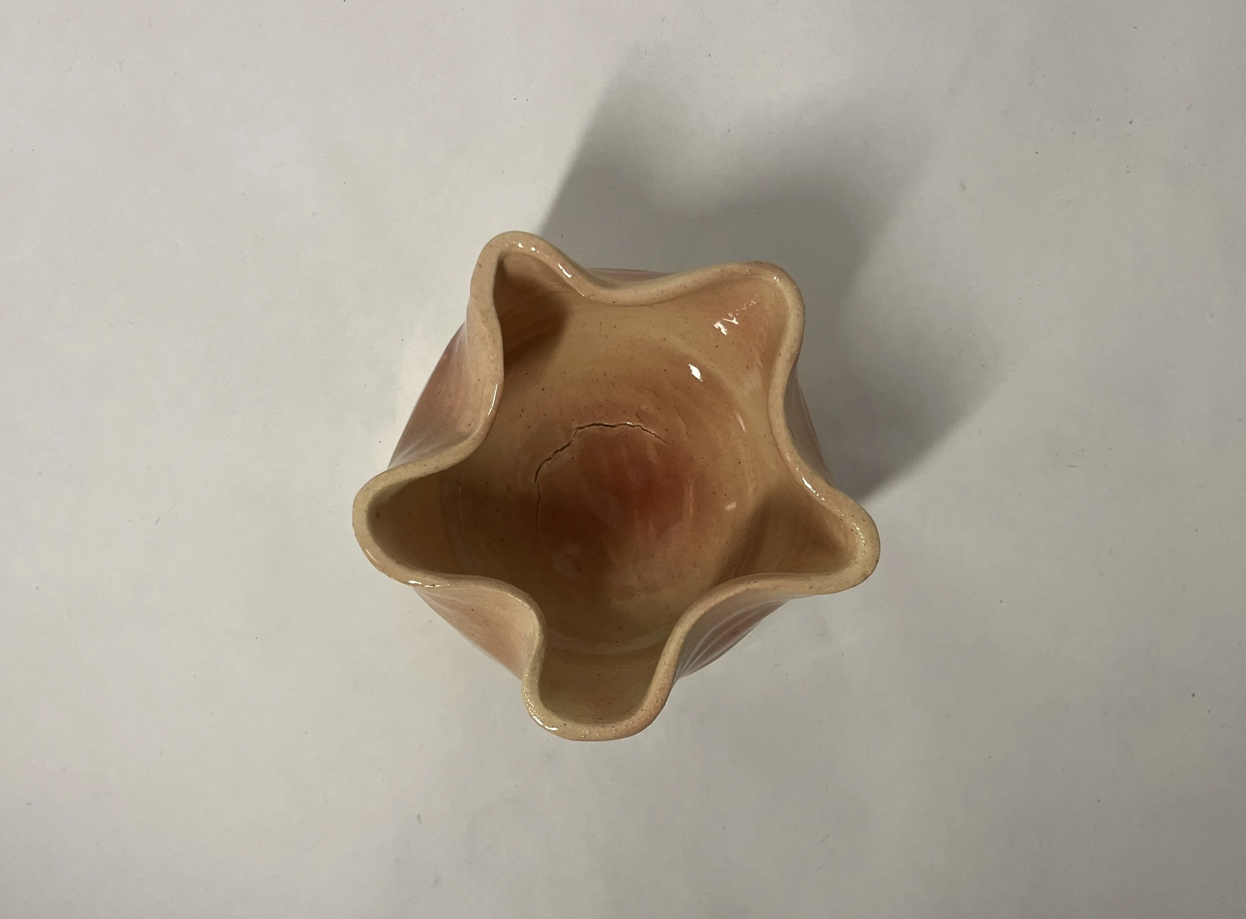 A ceramic vase with a ruffled, wavy rim, featuring a marbled beige and reddish-brown glaze, with a visible crack inside, resting on a white surface.