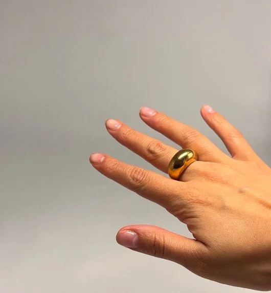 A hand wearing a large gold ring on the middle finger, with a plain, light-colored background.