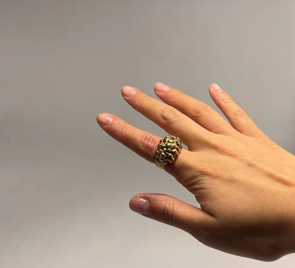 A person's hand wearing a large gold ring with a textured, cluster design, showing the back of the hand against a plain, light-colored background.