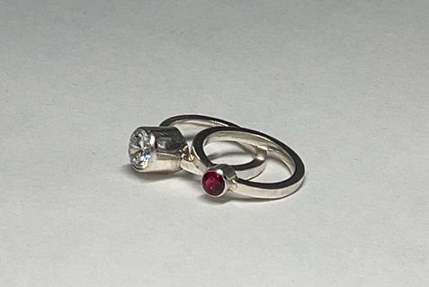 Two silver rings with gemstones, one with a clear gemstone and the other with a red gemstone, placed on a white surface.