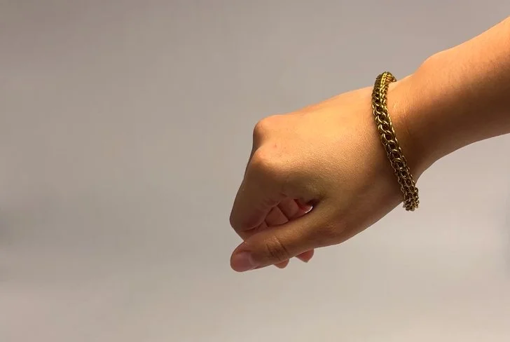A hand wearing a gold bracelet, clenched into a fist, against a plain gray background.