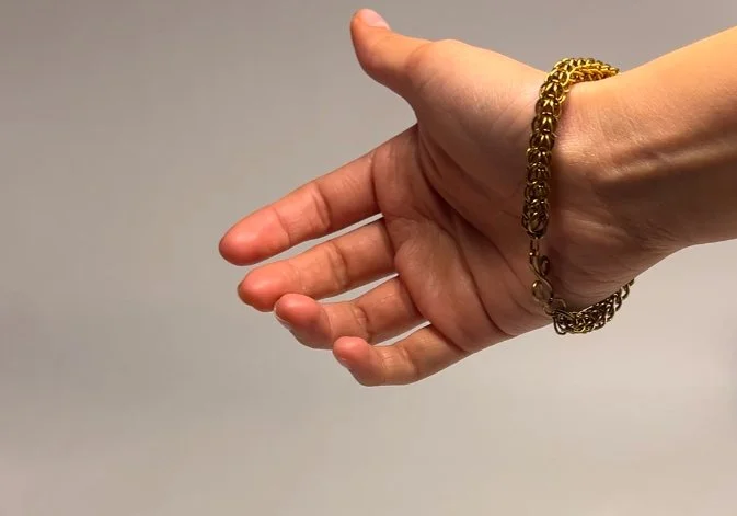 A person’s left hand extended with the palm facing down, wearing a gold bracelet, against a plain gray background.