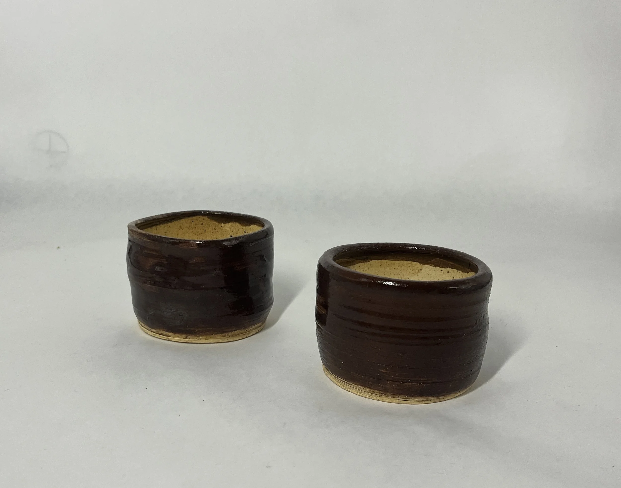 Two small brown ceramic cups with beige interior, placed on a white surface with a plain white background.