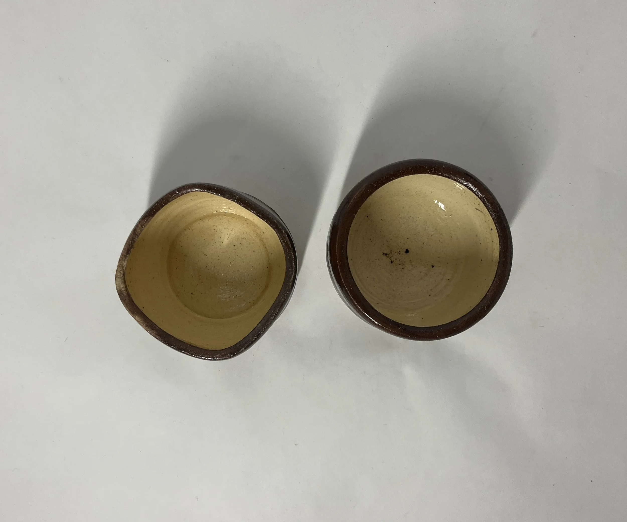 Two small, brown ceramic cups with beige interiors viewed from above, casting shadows on a white surface.