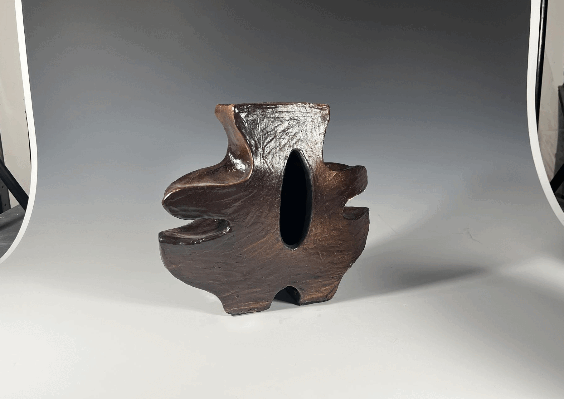 A wooden sculpture resembling a stylized animal with an oval hole in the center, set on a gray surface against a plain gray background.