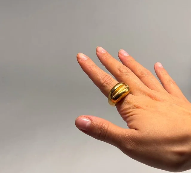 A person's hand with a large gold ring on the middle finger, against a plain light gray background.