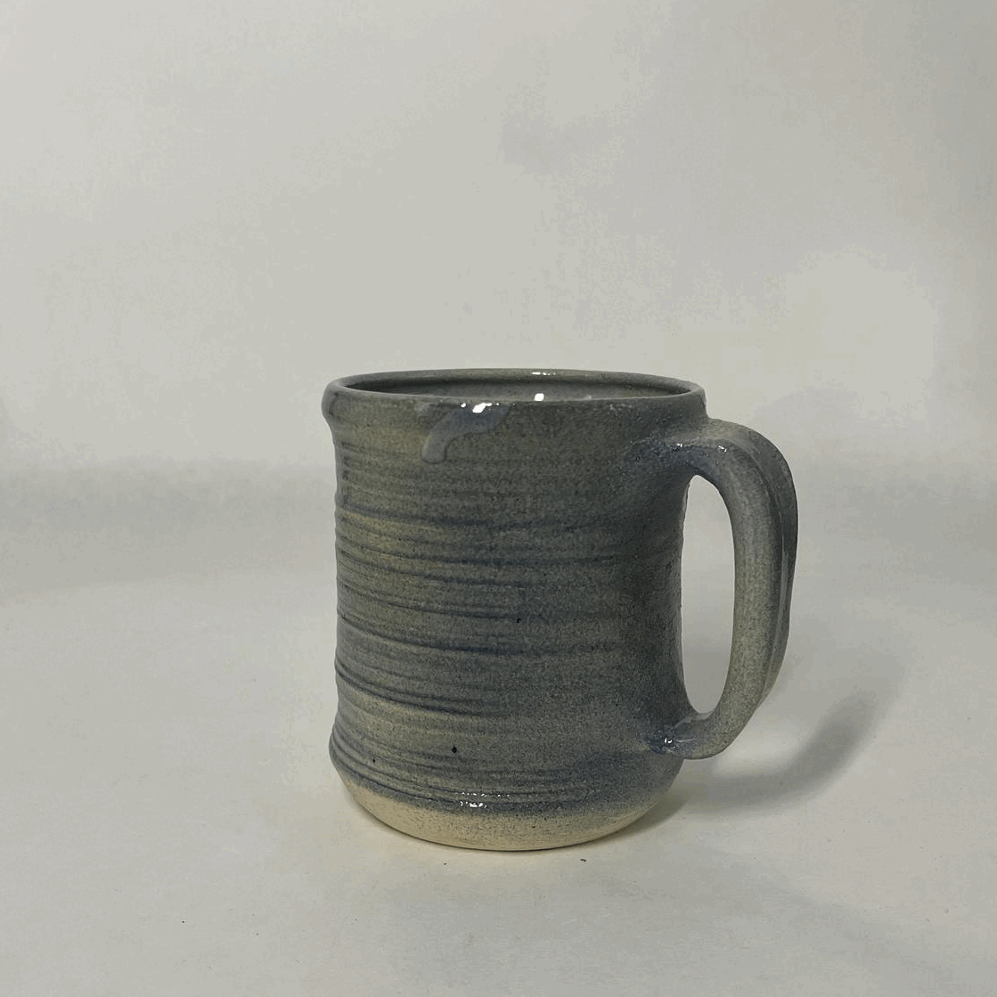 A ceramic mug with a gray and beige glazed finish, round shape, and a large handle placed on a plain white surface.