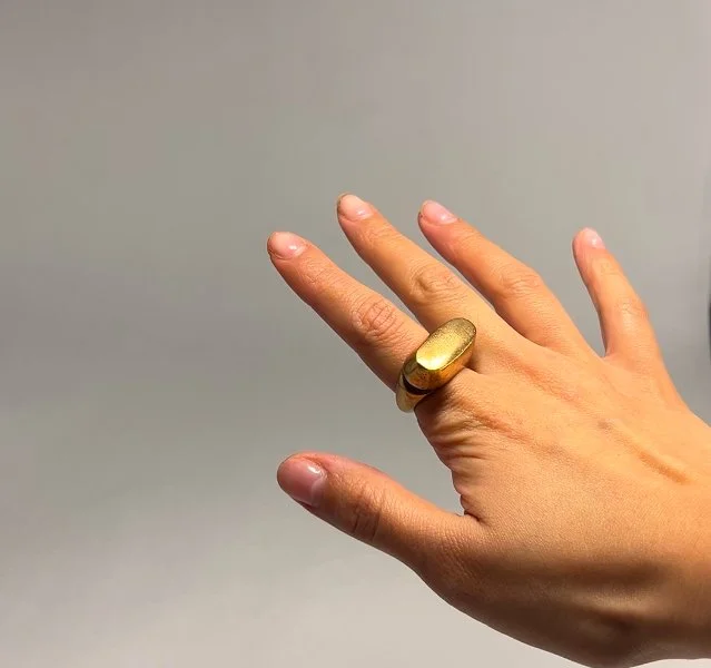 A hand with a gold ring on the index finger and middle finger, against a plain gray background.