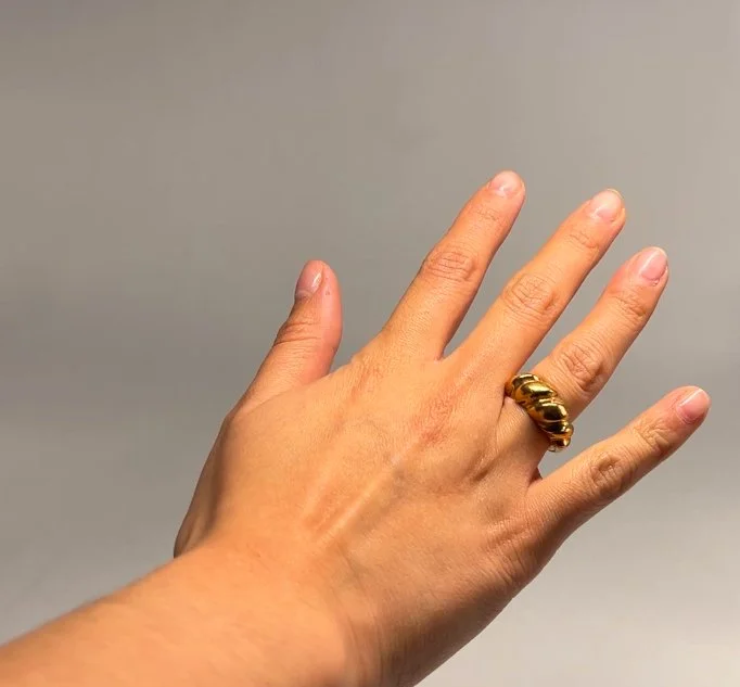 A person's left hand wearing a gold ring with a thick, rounded design on the ring finger, against a plain gray background.