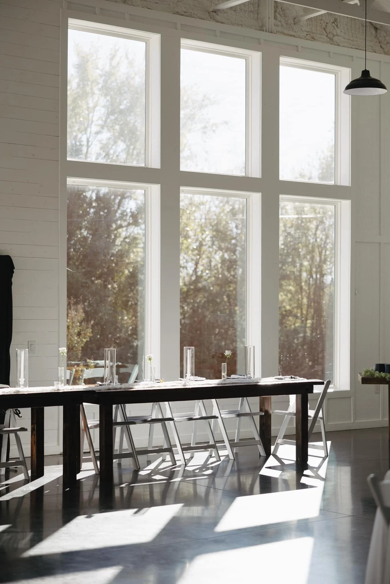 A long wooden table with white chairs and glass candlesticks arranged on top, set in front of large windows letting in natural sunlight, in a bright, minimalist room.