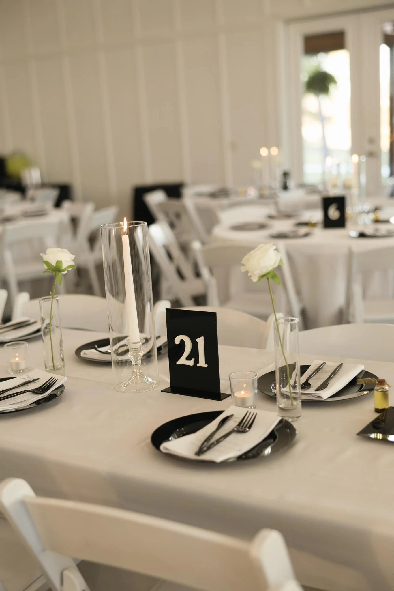 Elegant wedding reception table setup with black and white accents, featuring a tall glass candle holder with a lit candle, small vases with white flowers, white tablecloths, napkins, and cutlery, and a table number 21 sign.