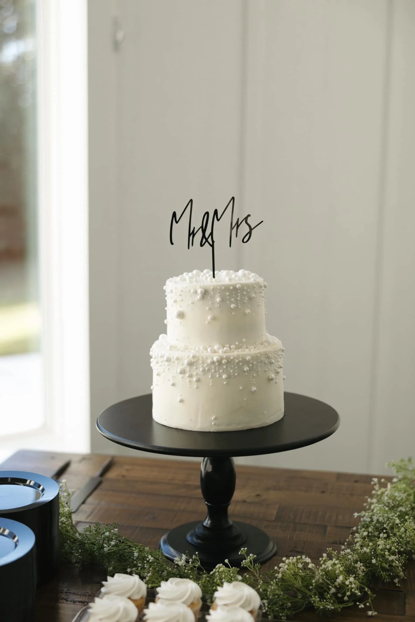 Two-tiered white wedding cake with pearl-like decorations on a black cake stand, topped with a black cake topper saying "Married" in cursive, surrounded by small white cupcakes and greenery.