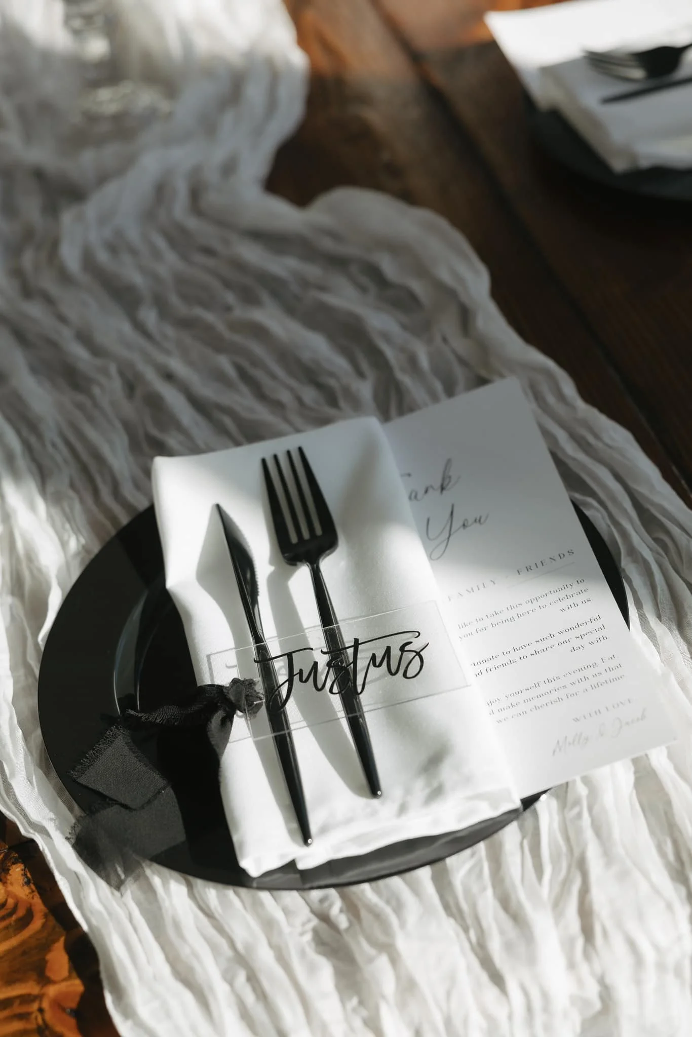 A table setting with a black plate, white napkin, black cutlery, a black ribbon, and a thank you note with elegant handwriting and a printed message, on a white textured tablecloth.