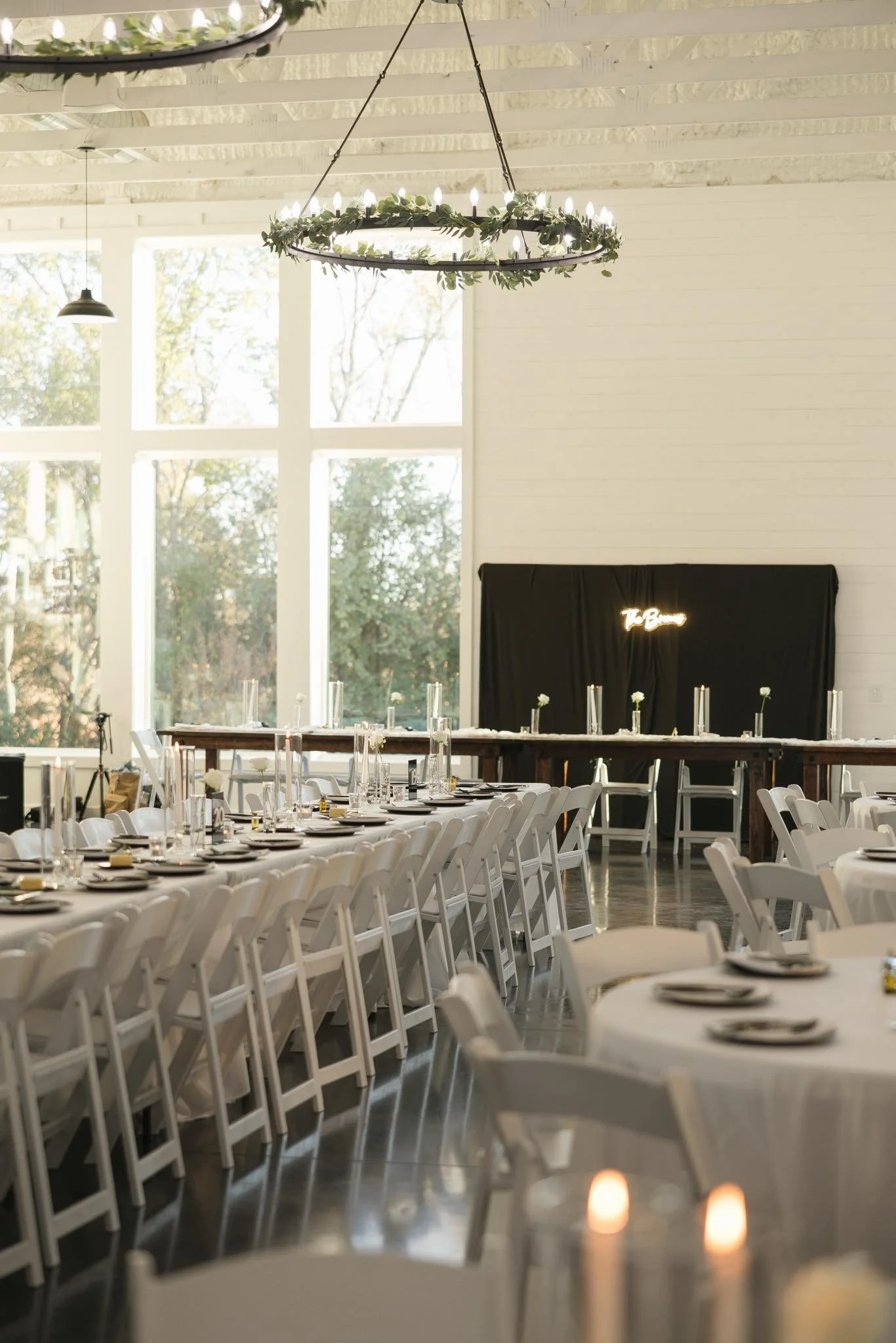 Elegant indoor event space with long tables covered in white tablecloths, decorated with plates, glasses, and tall candles. Modern chandelier and large windows with trees outside, black backdrop with neon sign reading 'The Boho' in the background.