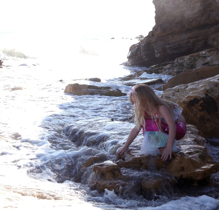 A young girl with long blonde hair wearing a colorful dress crouches on rocks by the ocean, touching the water as waves wash over the rocks near a rock formation.