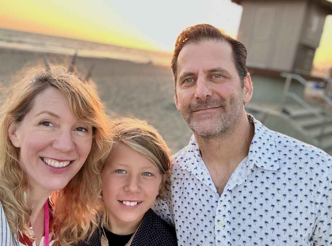 A family of three taking a selfie at the beach during sunset. Woman with blonde hair, man with dark hair and beard, and young boy with blonde hair, all smiling.