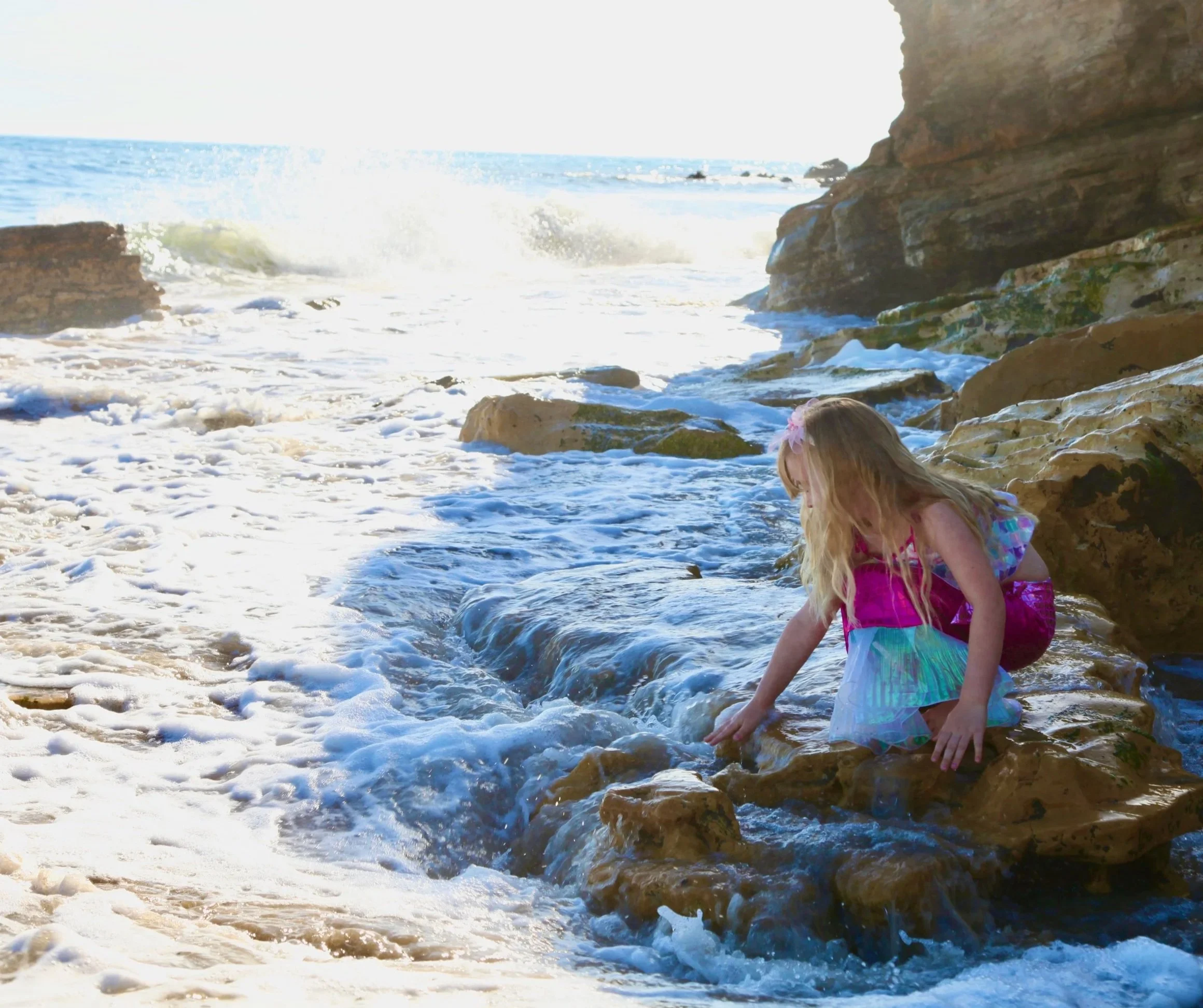 A young girl in a colorful dress playing in the ocean waves on a rocky beach, with cliffs in the background on a bright sunny day.