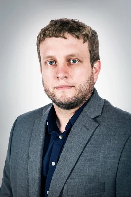 Headshot of a man with short brown hair and blue eyes, wearing a gray blazer and a dark blue shirt, against a plain light gray background.