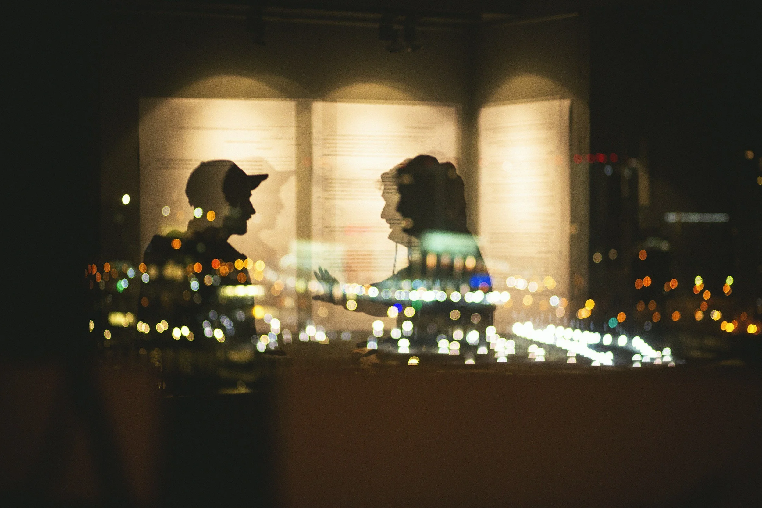 Nighttime scene of a book with silhouettes of two people inside, reflected on a window with city lights blurred in the background.