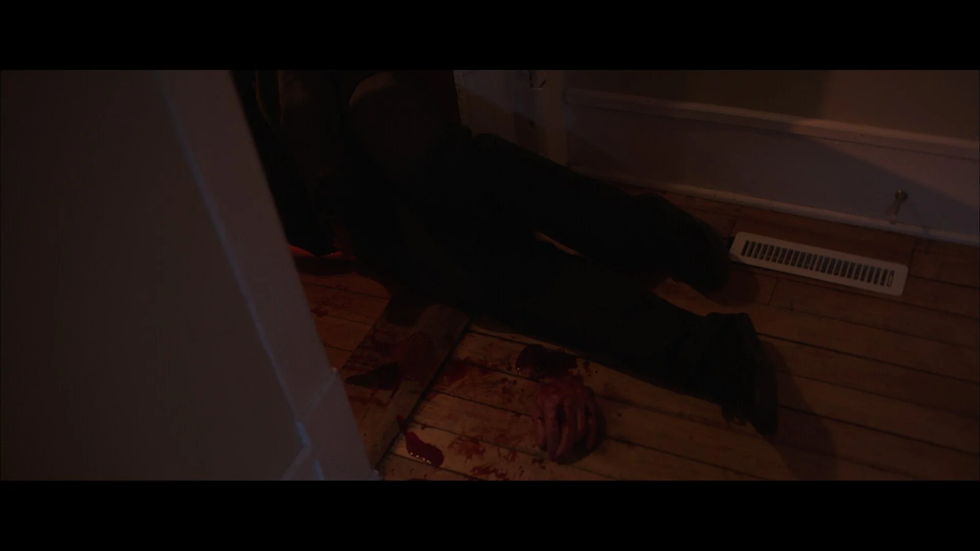 A person lying on the wooden floor with bloodstains, with its face not visible, near a wall and a door, with some blood drops and a bloodied cloth nearby.