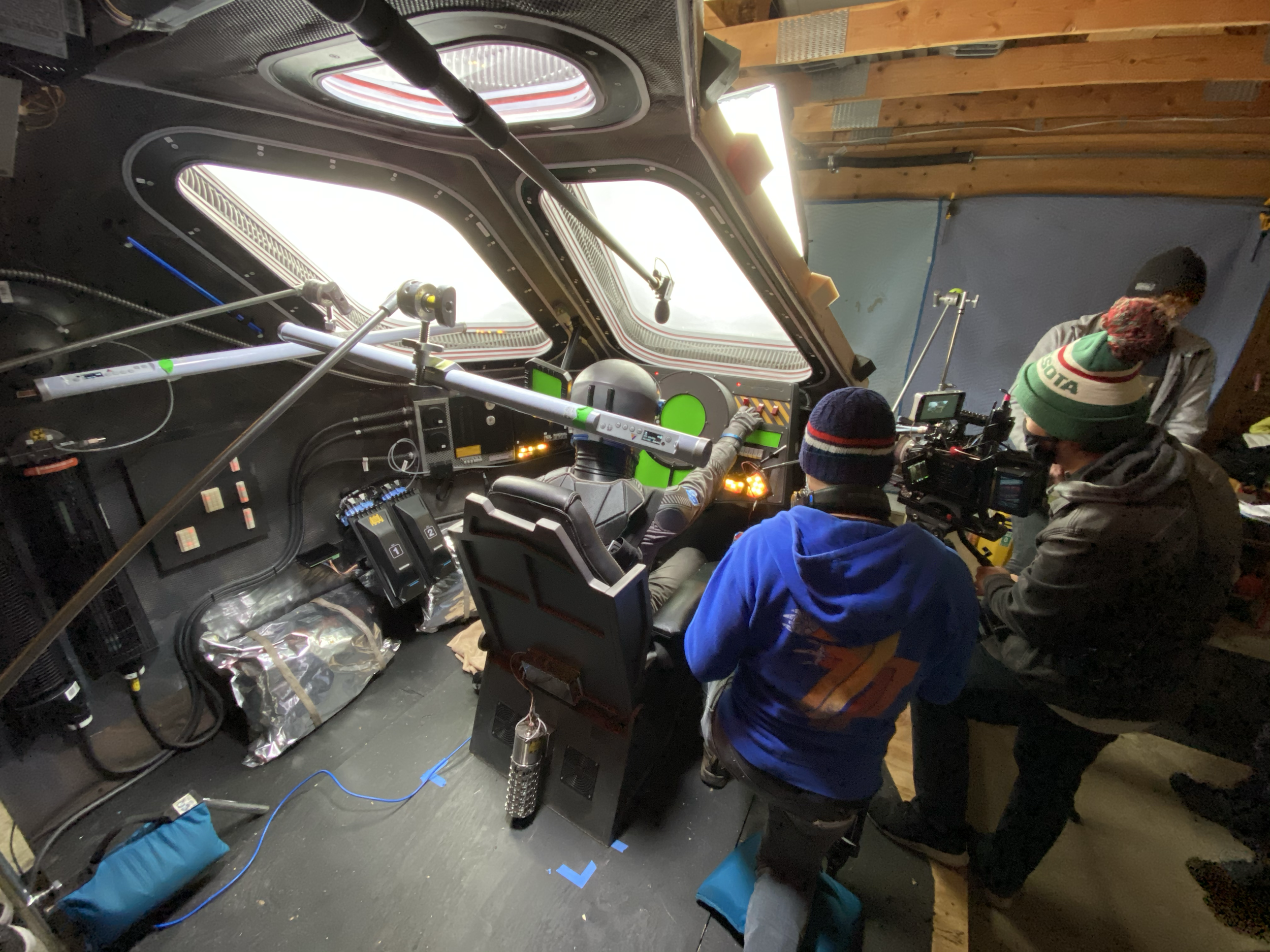 Inside a spaceship or aircraft cockpit with multiple electronic screens and controls, and people wearing winter clothing, including a green hat with the word 'SOTA' on it, engaged in a discussion or activity.
