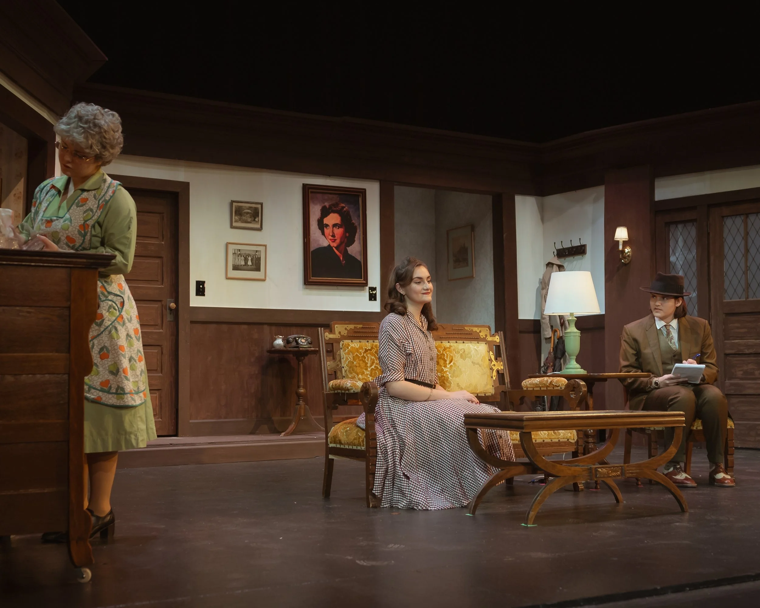 A scene from a theatrical play with four actors dressed in vintage clothing sitting in a cozy room with wood-paneled walls, framed pictures, and warm lighting.