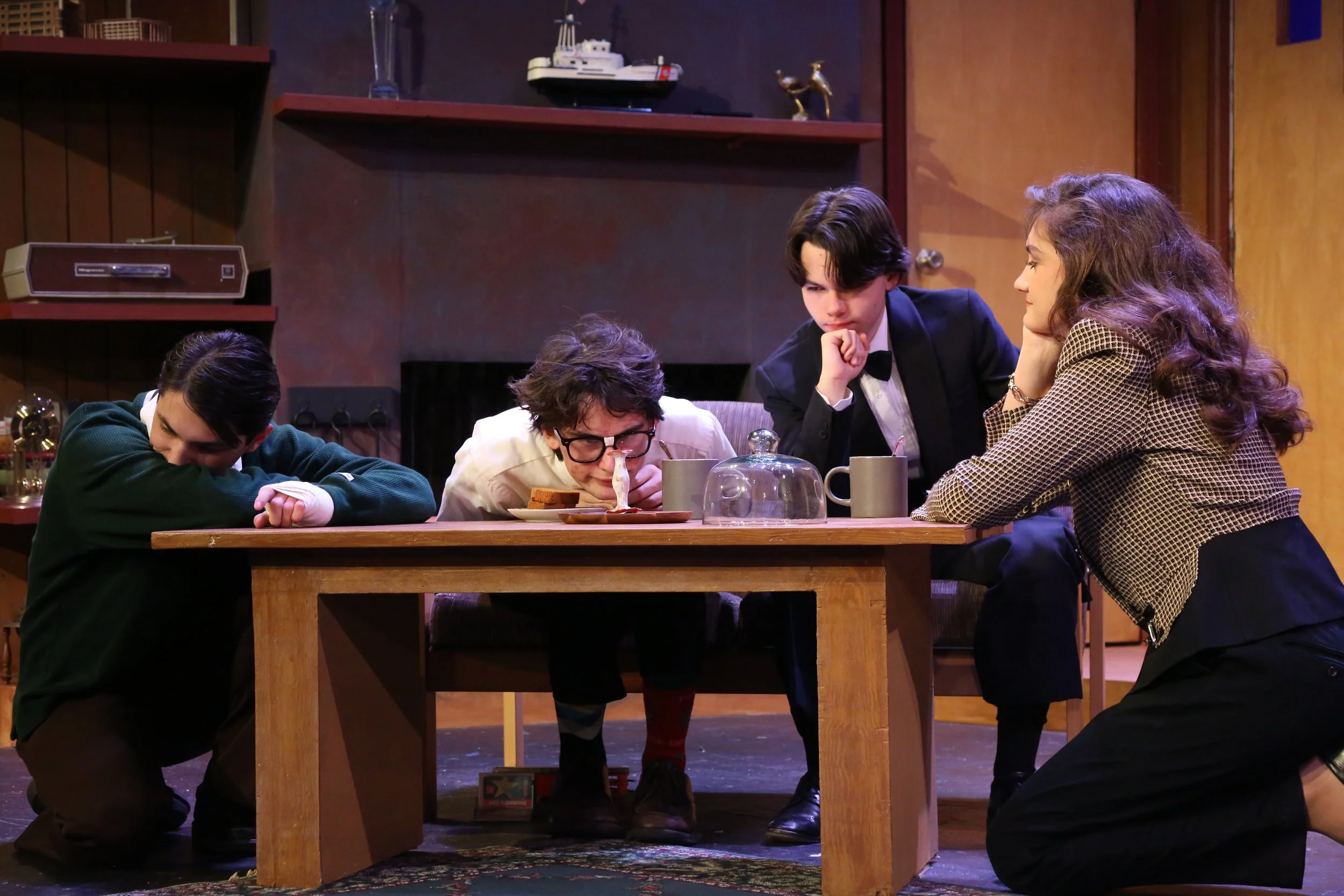 Four young actors in costume performing a scene on stage with a wooden table in a setting resembling a dining room.