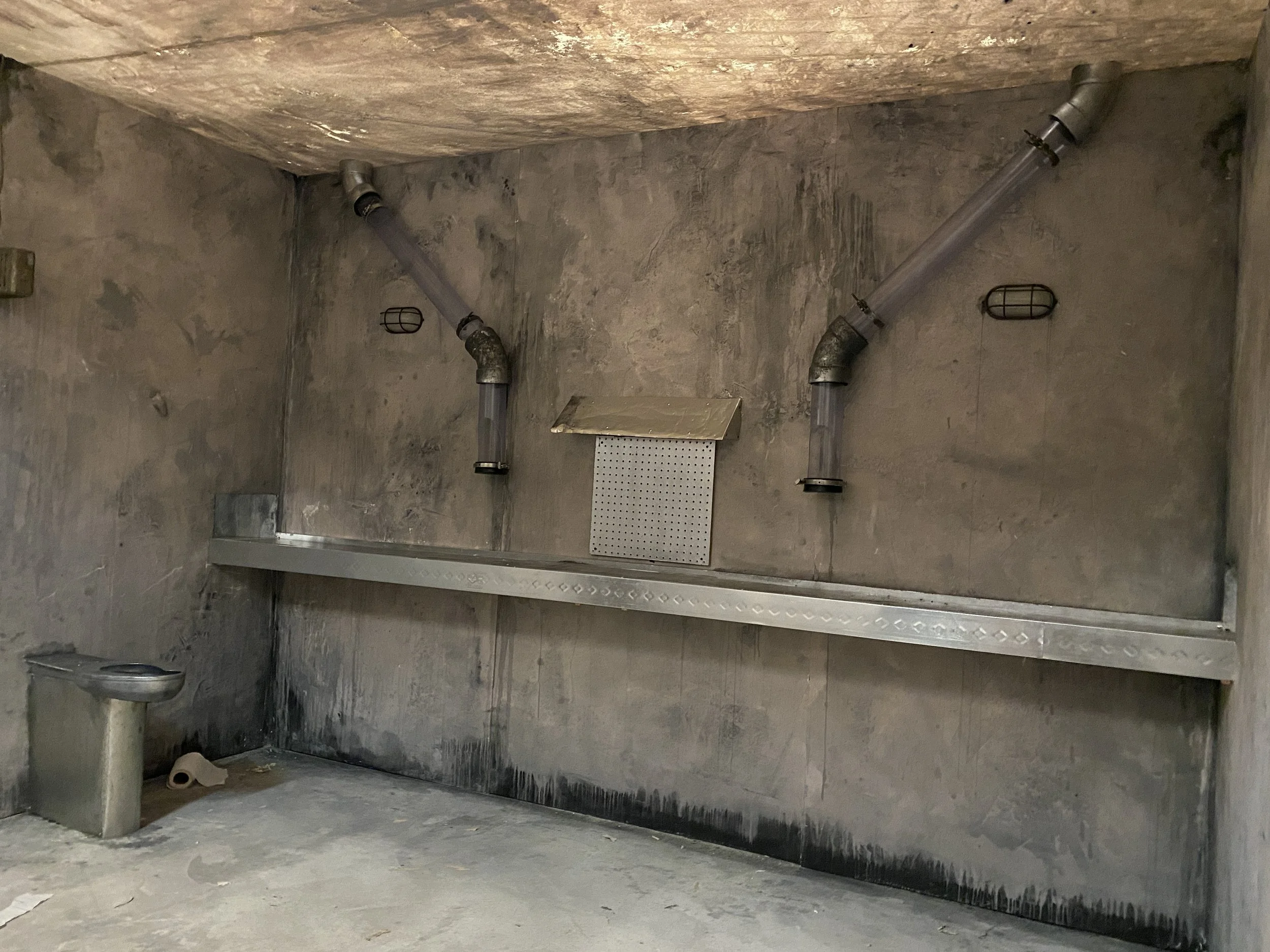 Film set of an industrial room with concrete walls and floor, featuring two metal pipes on the ceiling, a small sink on the left, and a metal drain cover on the floor.