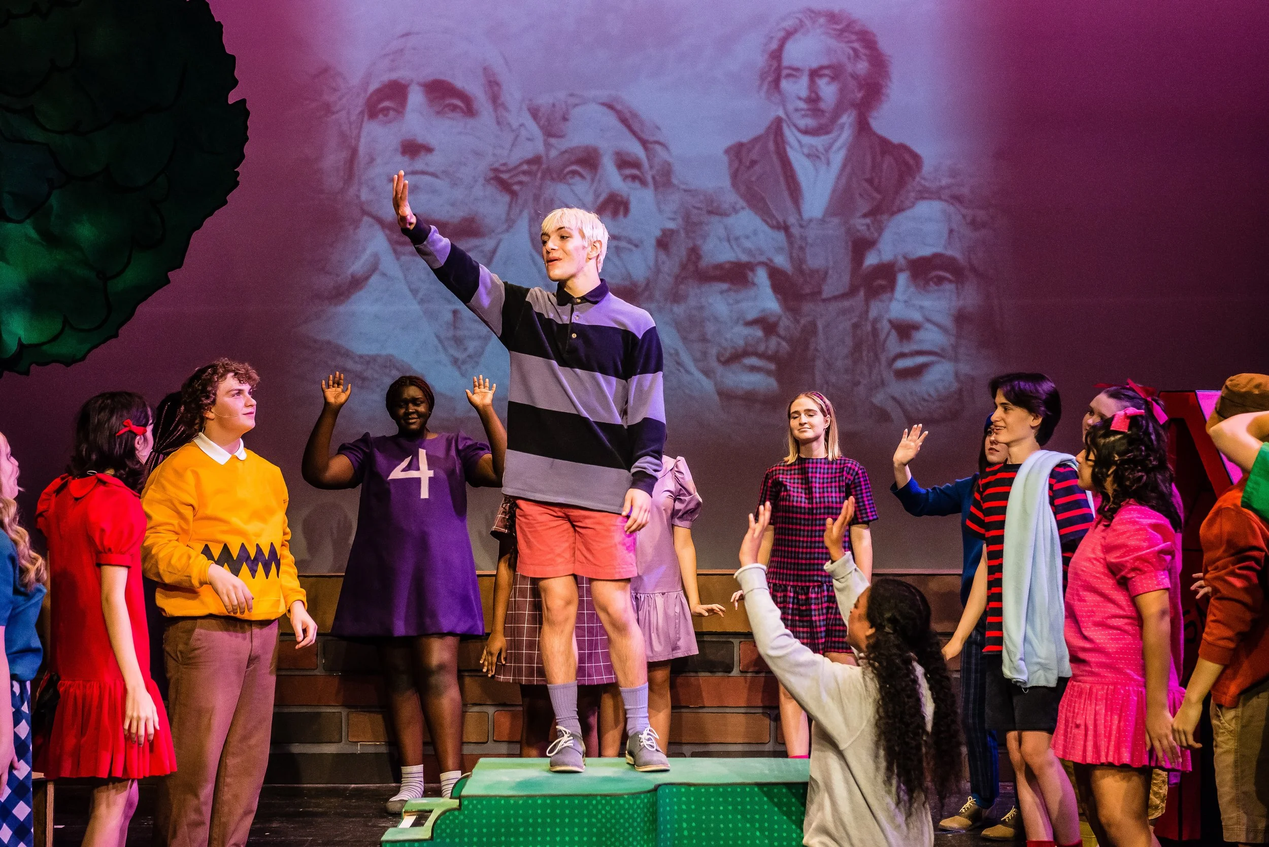 A stage performance with diverse children and teenagers, some raising their hands. A young person with short blond hair is standing on a green platform at the center, with a backdrop featuring Mt. Rushmore.