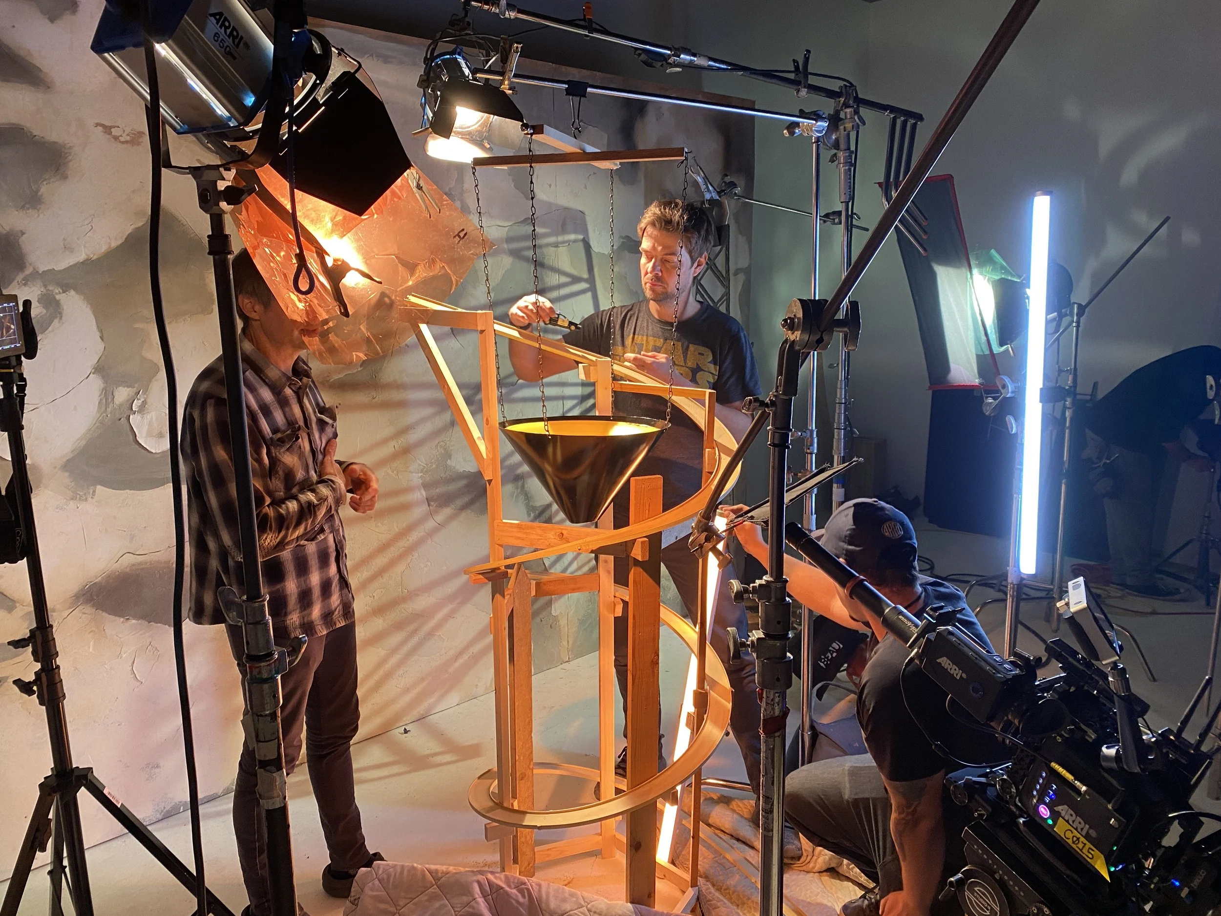 People working on a film effect model with bright lighting and film equipment around. A man with a beard and t-shirt is holding a tool, another person is observing, and there are cameras and lights set up for filming.