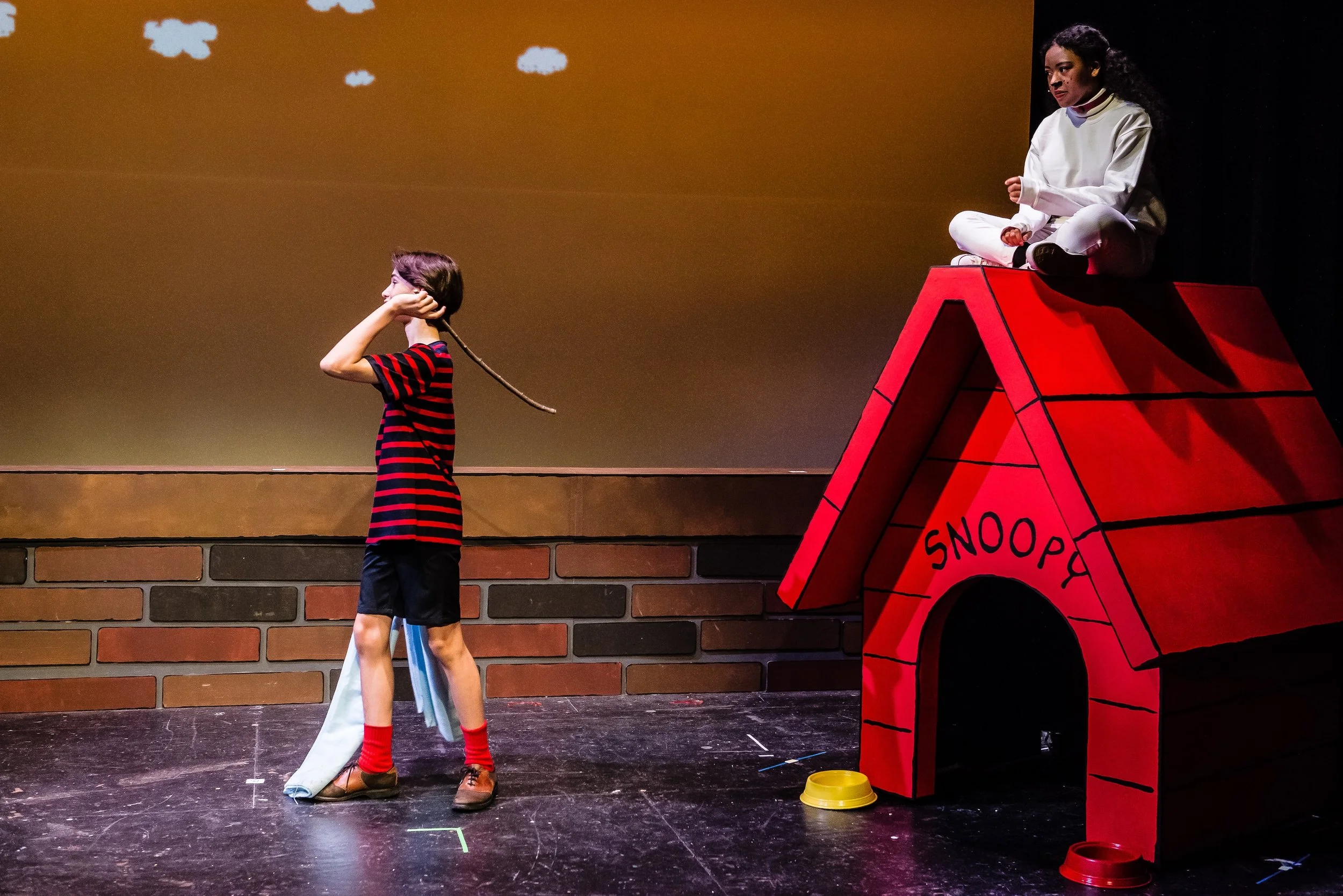 Two young actors on stage, one standing and the other sitting on a red doghouse with the word 'SNOOPY' written on it, against a backdrop of a brown wall with clouds in the sky.