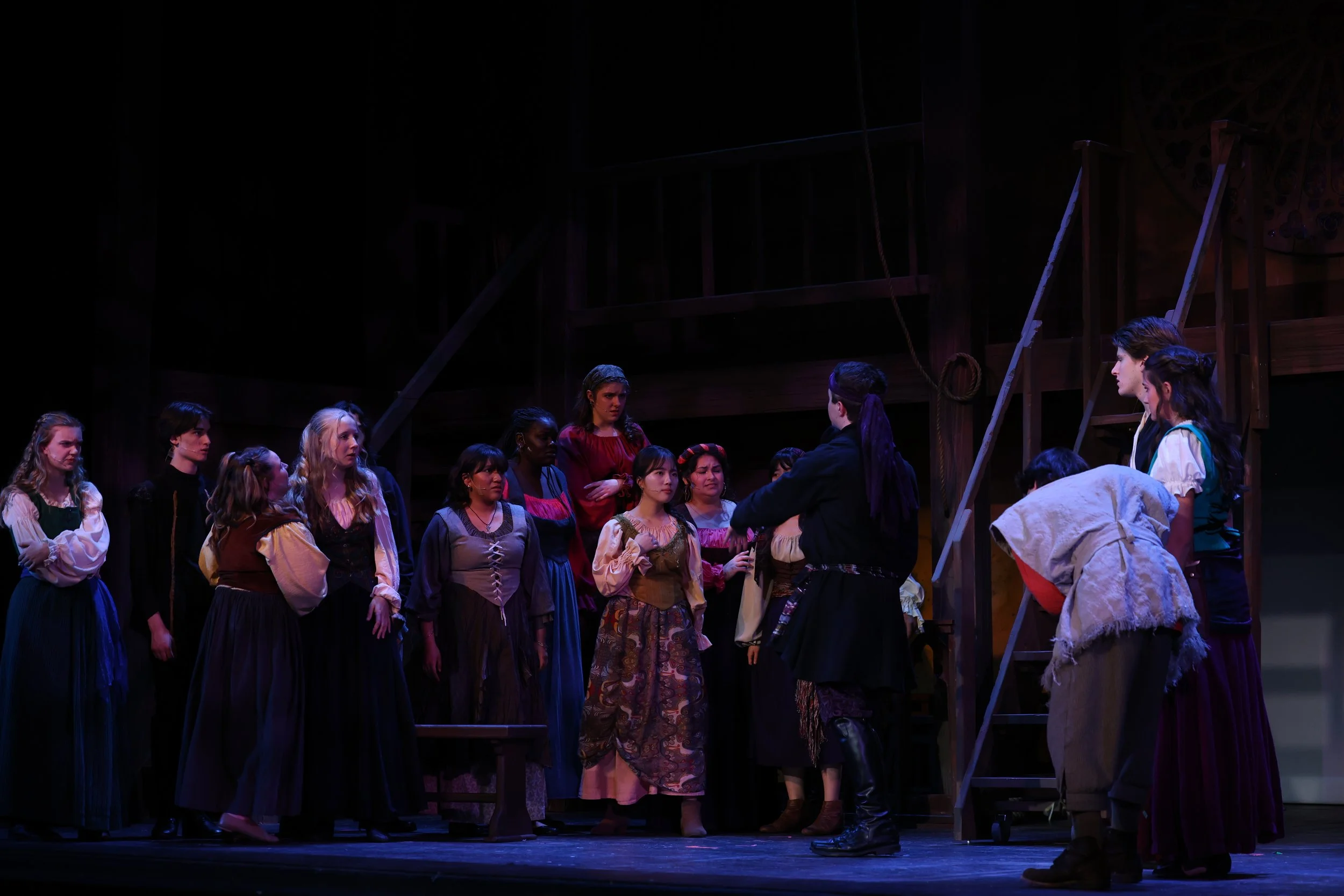 A group of people in costume on a theater stage, with some standing and others kneeling, engaged in a performance. The stage has a dark background with wooden structures and ladders.