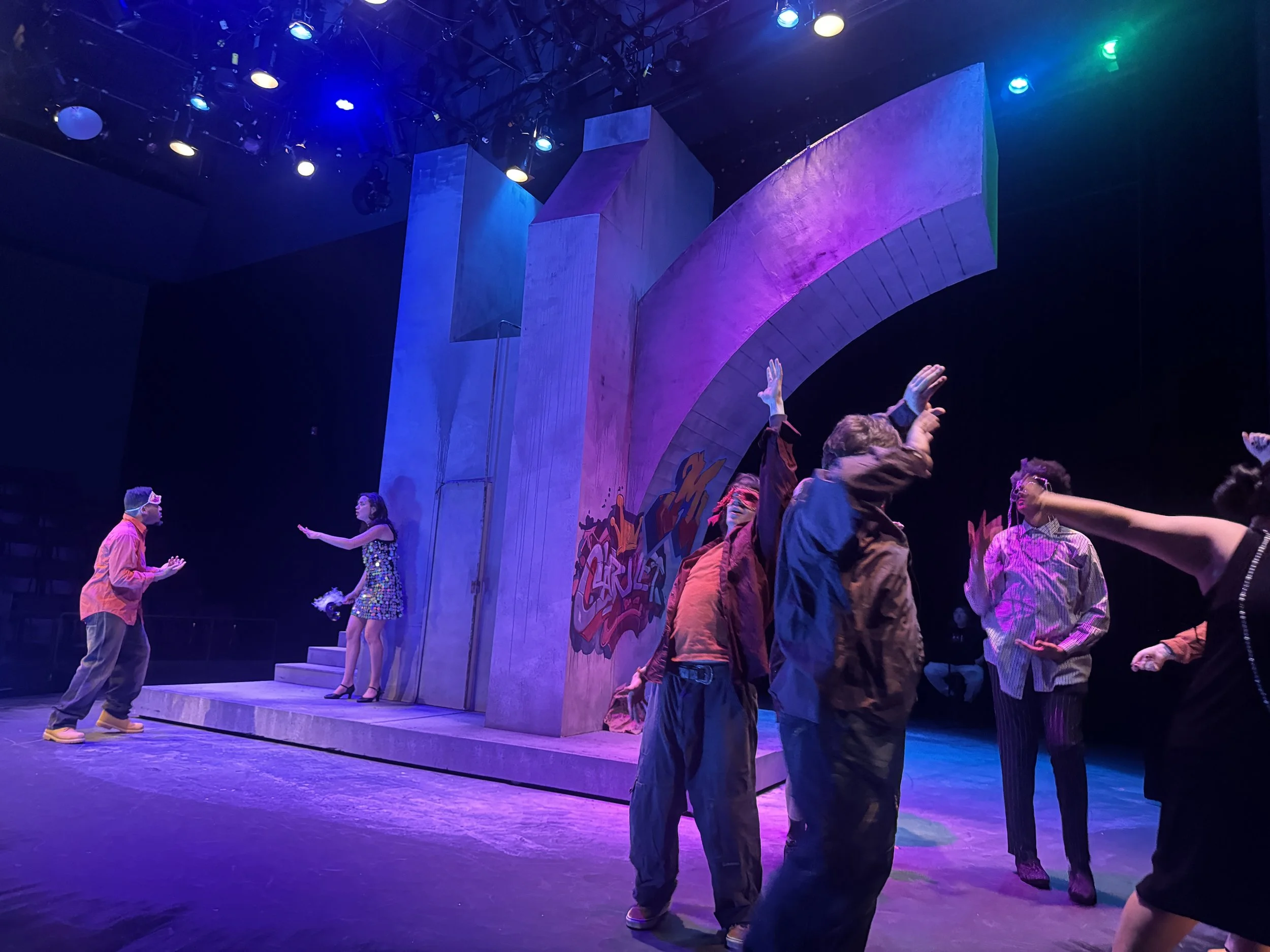 People performing on a stage with colorful lighting and a set design resembling a large painted concrete structure.