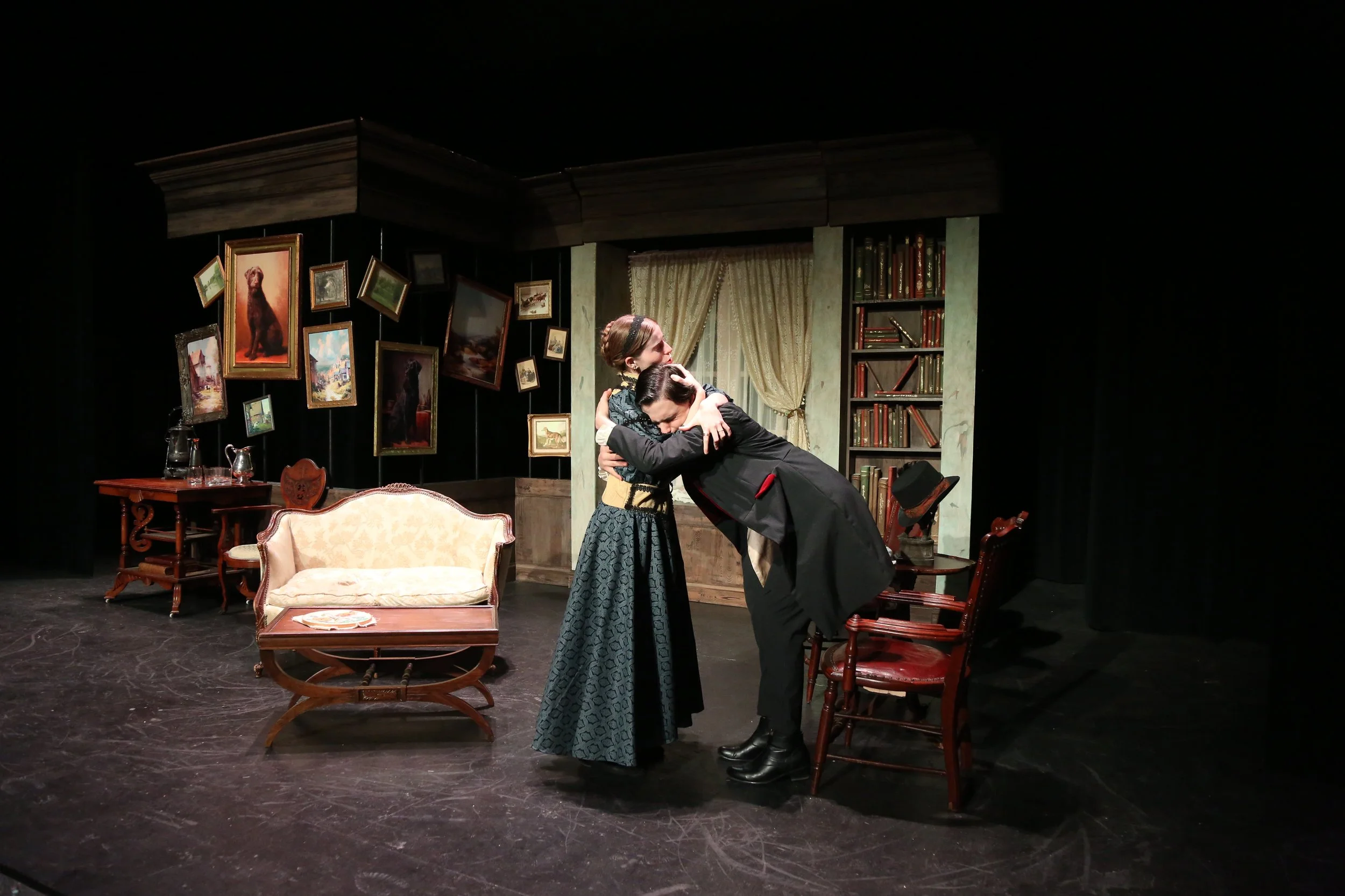 Stage scene of two actors, a man and a woman, embracing in a vintage room with framed paintings, a cream-colored sofa, a wooden coffee table, and bookshelves.