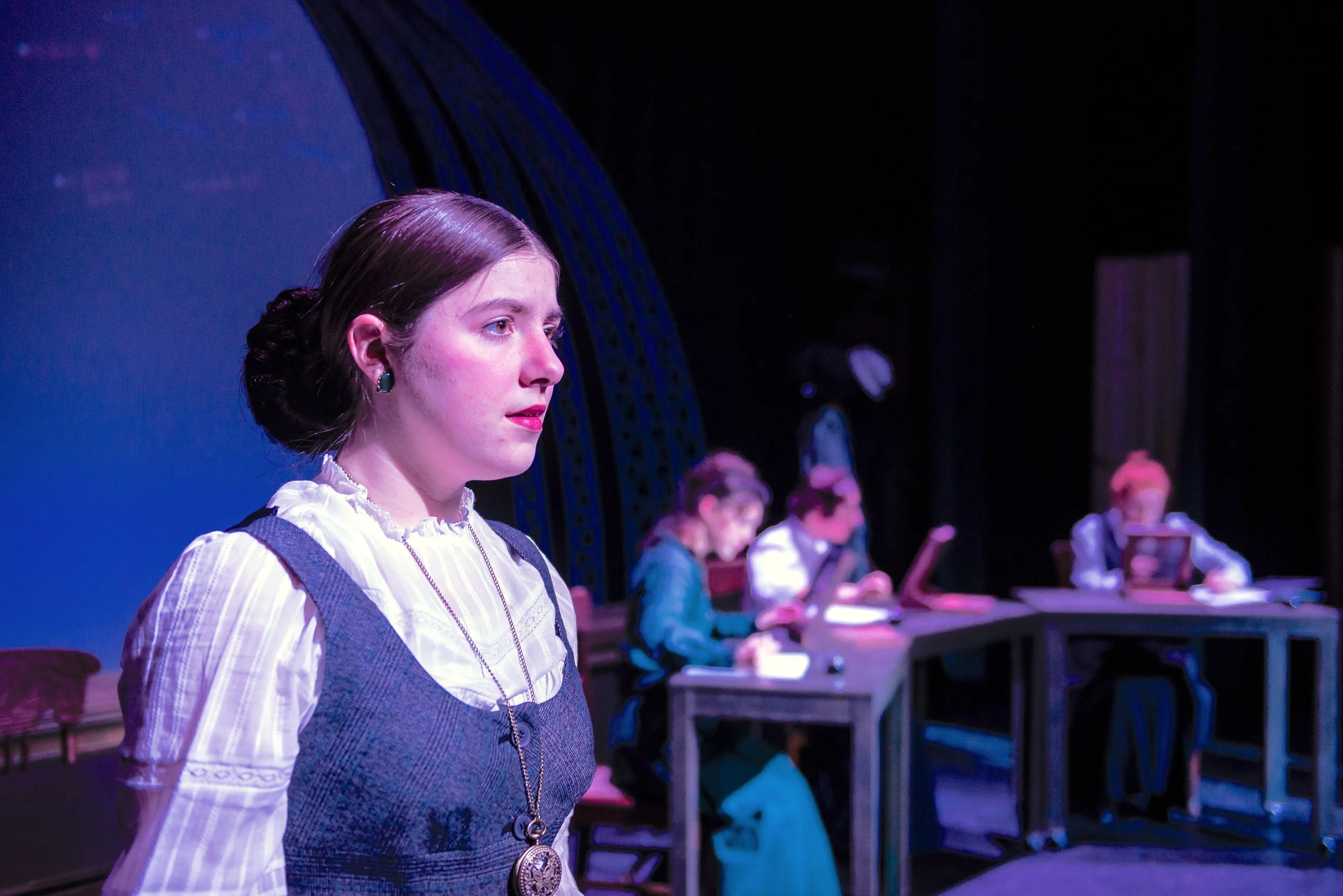 A young woman with brown hair styled in a bun, wearing a white blouse, a gray vest, and a large medallion necklace, stands in profile on stage with a serious expression. In the background, four other women sit at a long table, engaged in reading or w