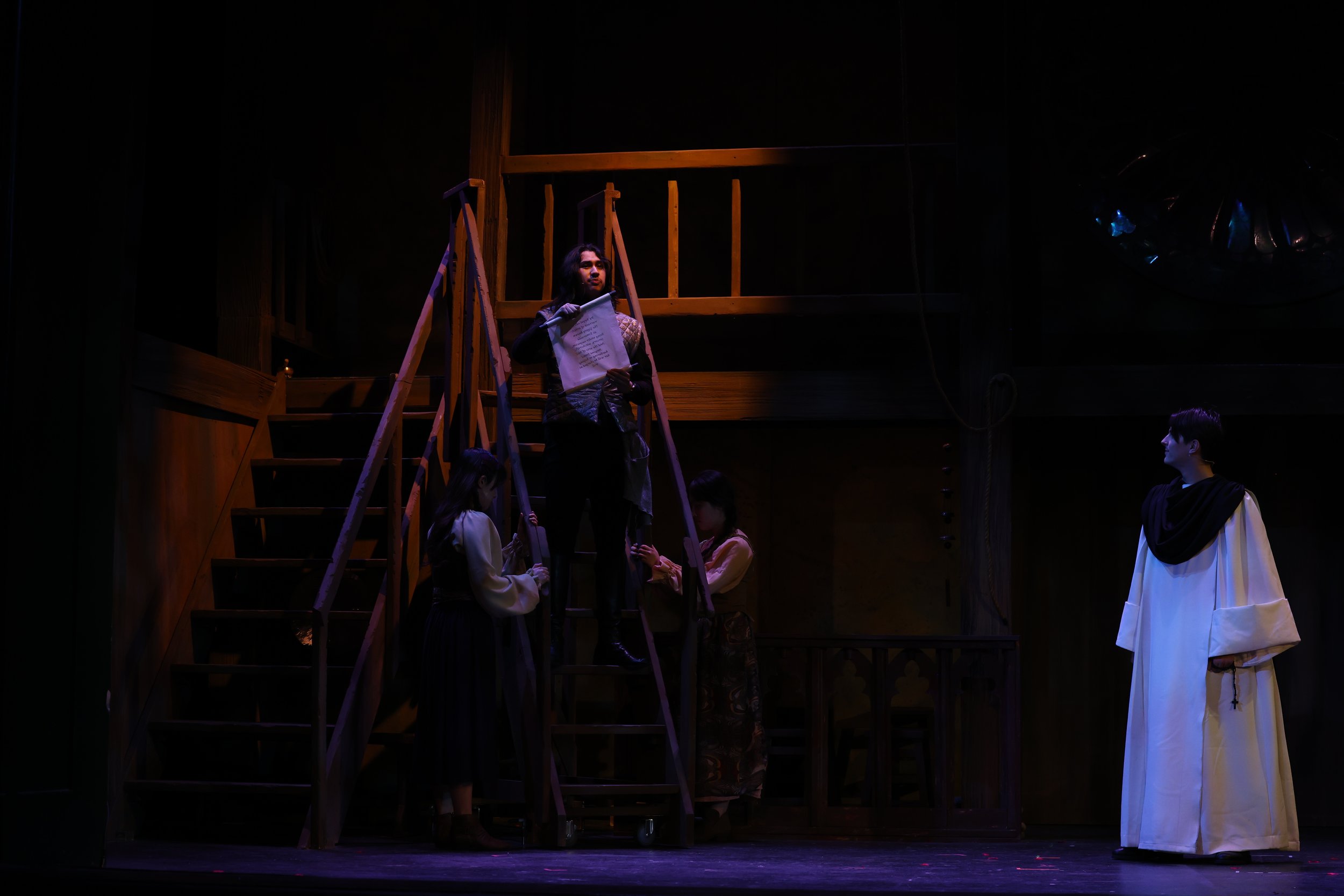 Stage scene with actors, one on a ladder holding a script, others gathered around, dark background, wooden set, partially lit.