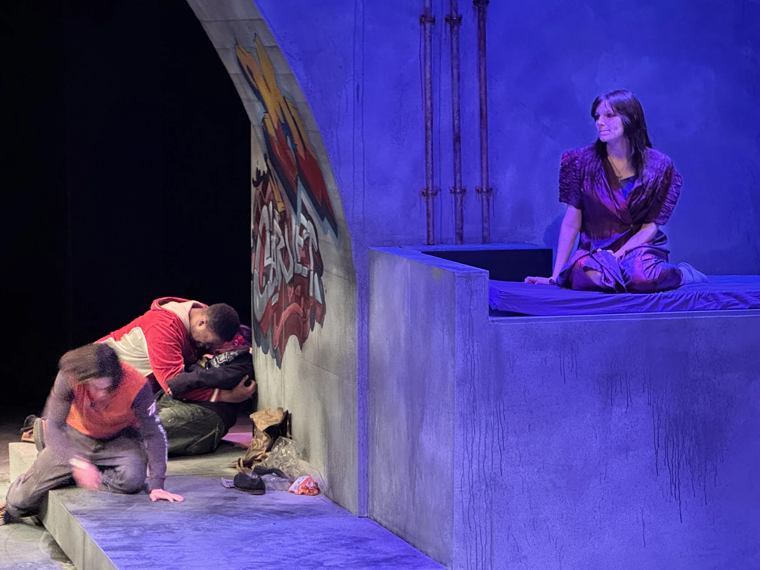 People sitting on a stage floor, with some backpacks and bags nearby, in front of a painted wall and a blue-lit background.