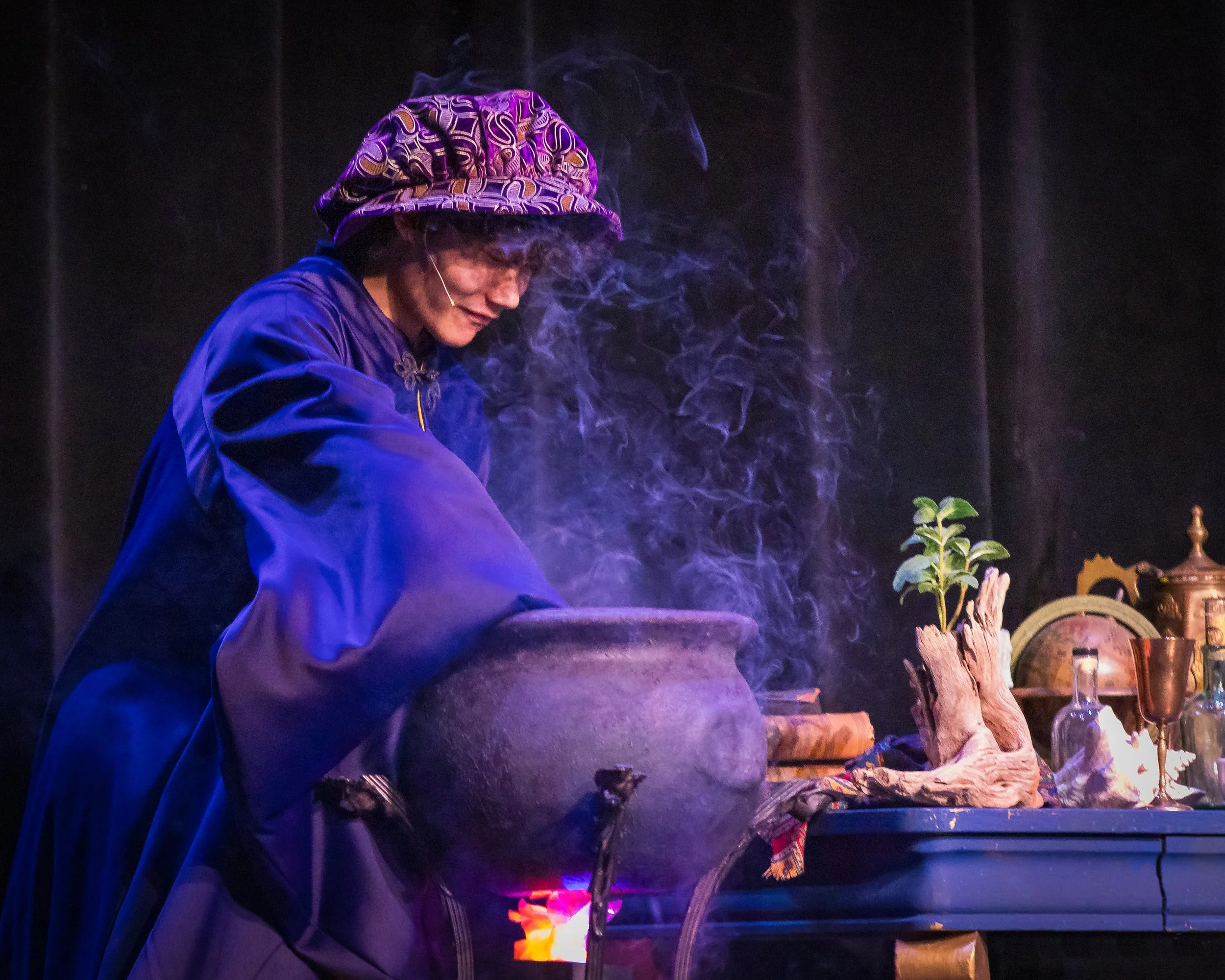 A person wearing a purple jacket and a colorful patterned hat is smiling and working with fire or flames in a dark setting. There are various decorative objects and plants on the table beside them, illuminated by the fire.