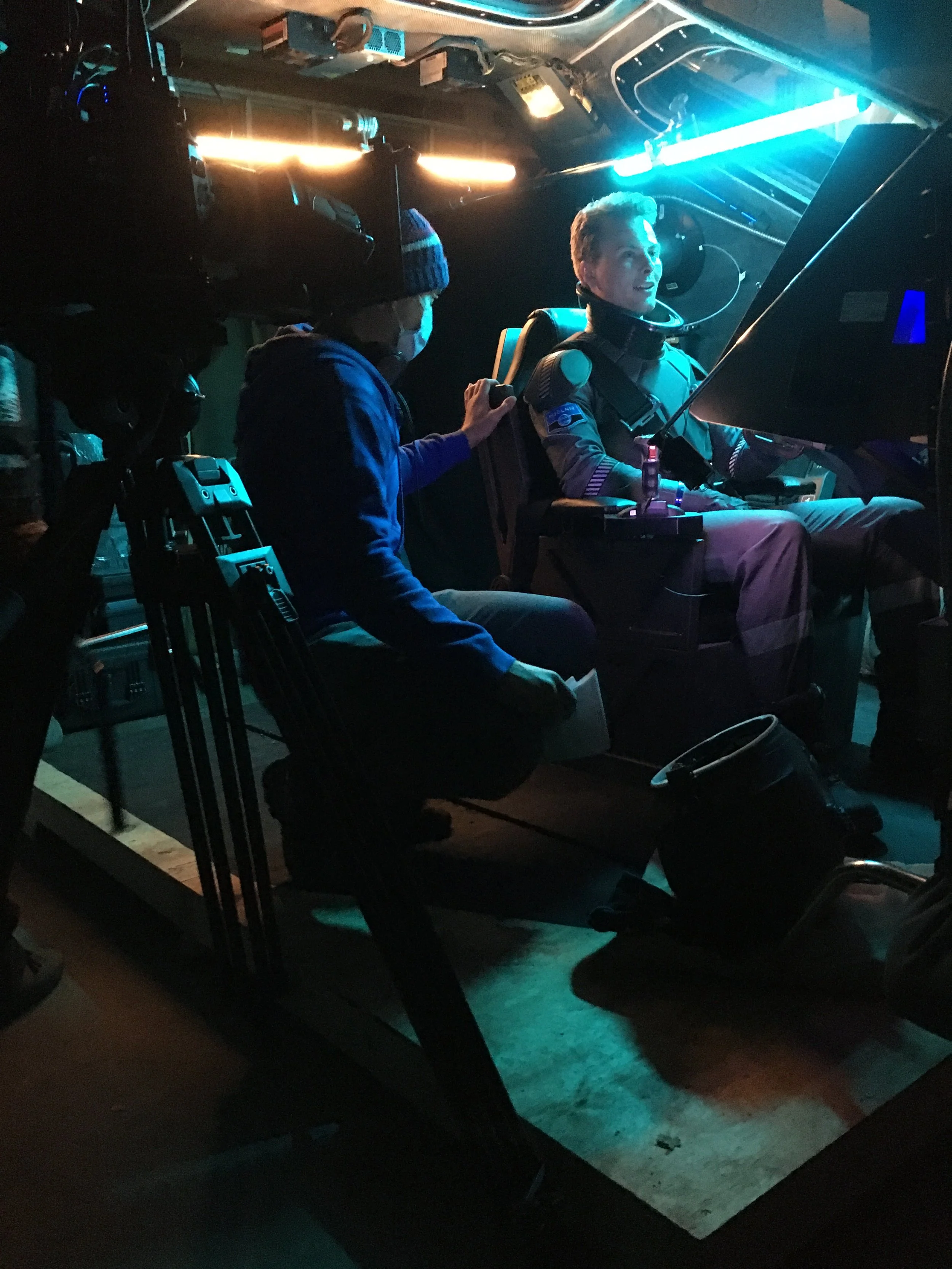 A person sitting in the set of a spacecraft cockpit with a spacesuit, talking to the director wearing a hat, surrounded by control panels and illuminated by blue and orange lights.