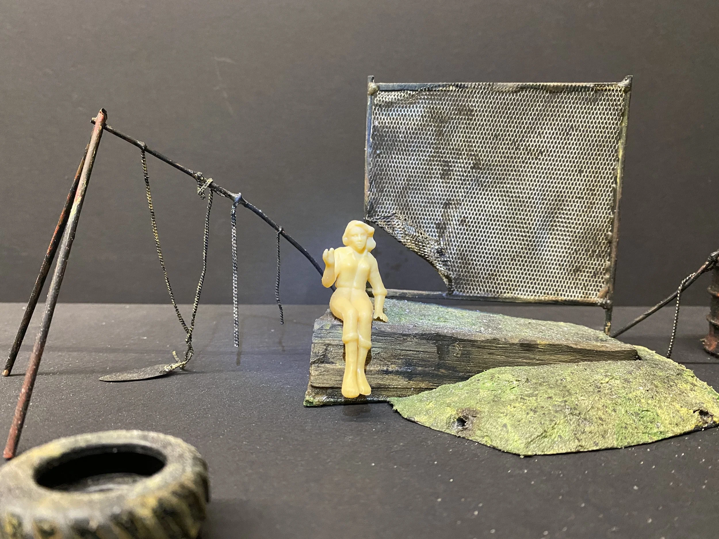 A miniature scene featuring a small figurine of a woman sitting on a wooden platform next to a miniature metal fence, with various small objects and leaves around on a black surface.