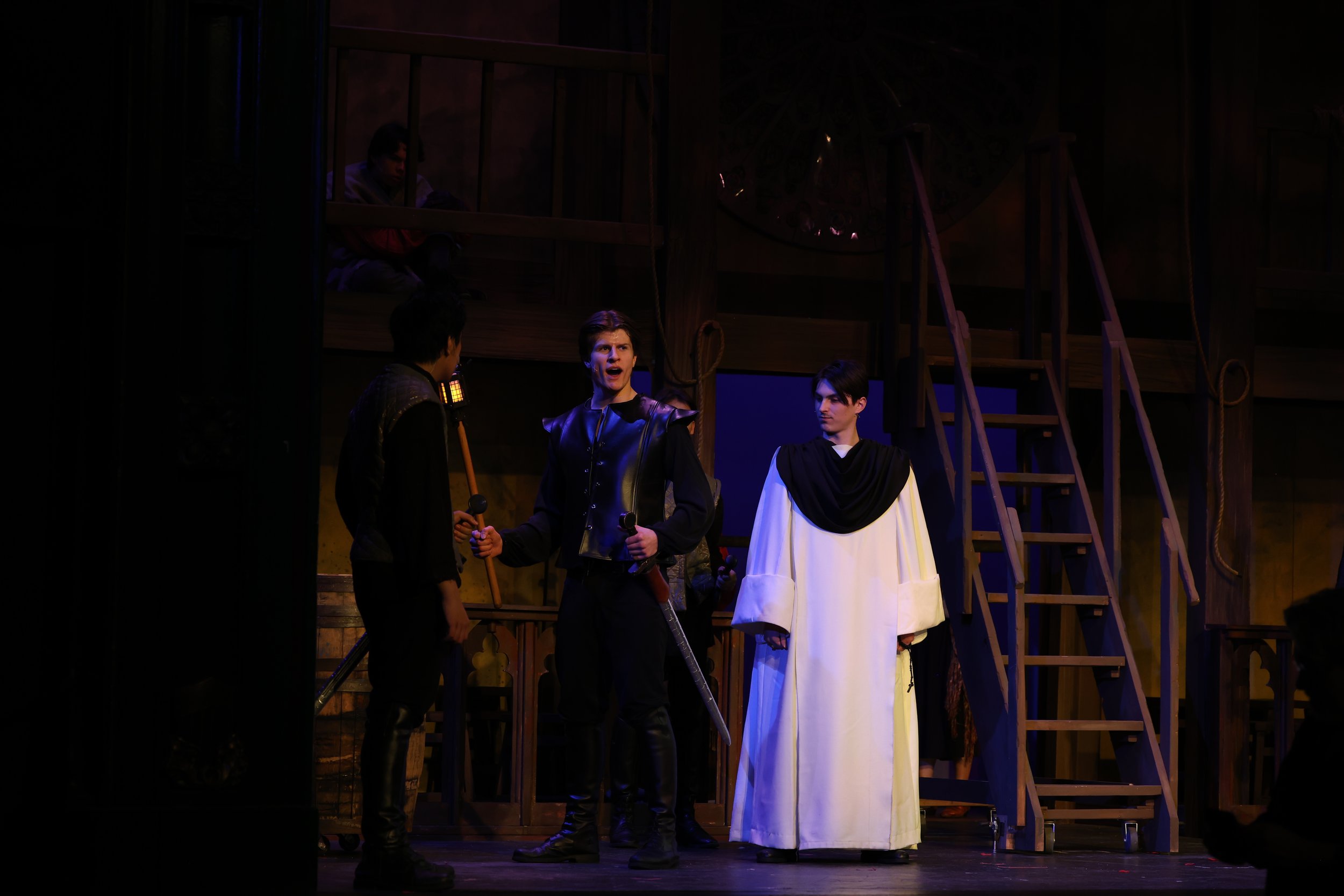 Three male actors in costumes performing on a theater stage with a wooden set and a person in the background observing.