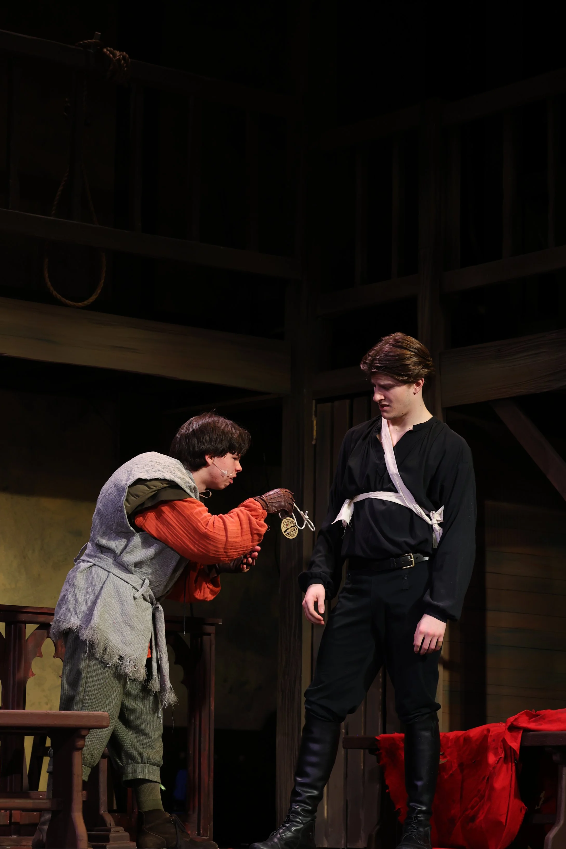 Two actors in a theatrical scene: one young man with dark hair, wearing a black costume, standing with a concerned expression; another young man with dark hair, dressed in gray and orange costume, kneeling and handing an object to the standing man, i