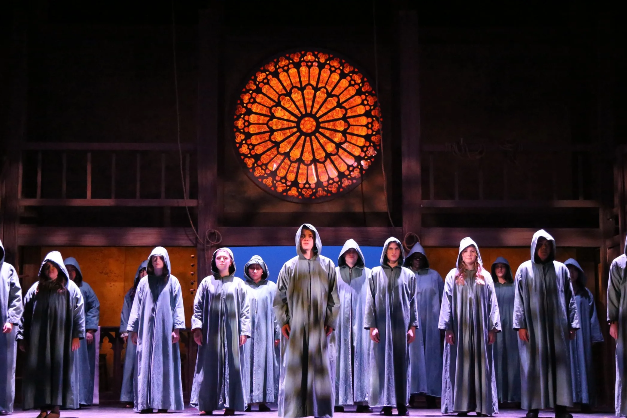 Group of actors dressed in gray robes with hoods standing in a church or cathedral with a large stained glass rose window in the background.