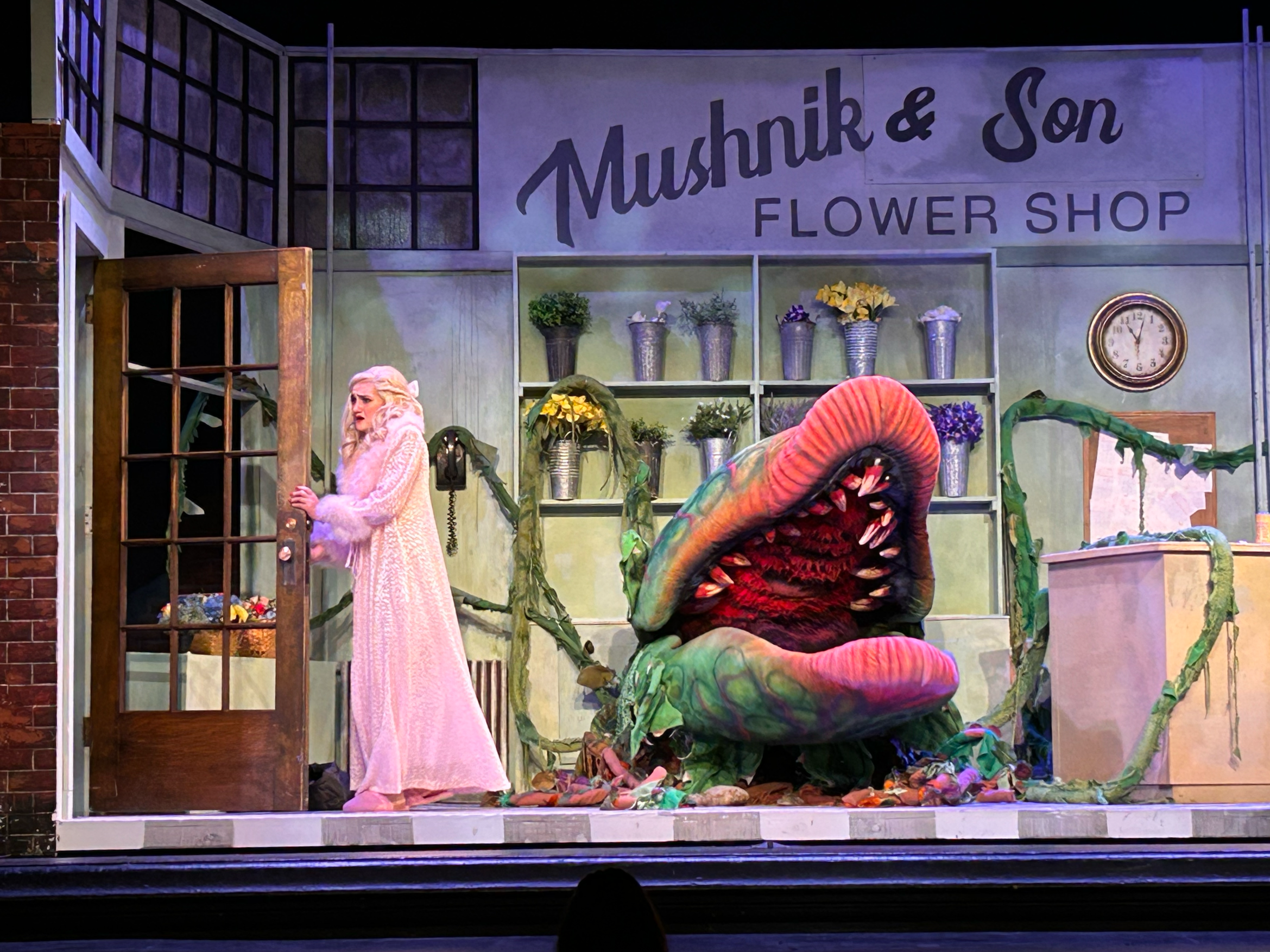 A stage set for a play with a flower shop sign, featuring a woman in a pink dress and a large colorful flesh-eating plant.