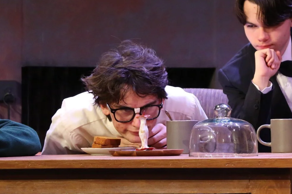 Two young men at a table, one with glasses leaning close to a plate, observing an apple core. The other man in a tuxedo looks thoughtful, with his hand on his chin.