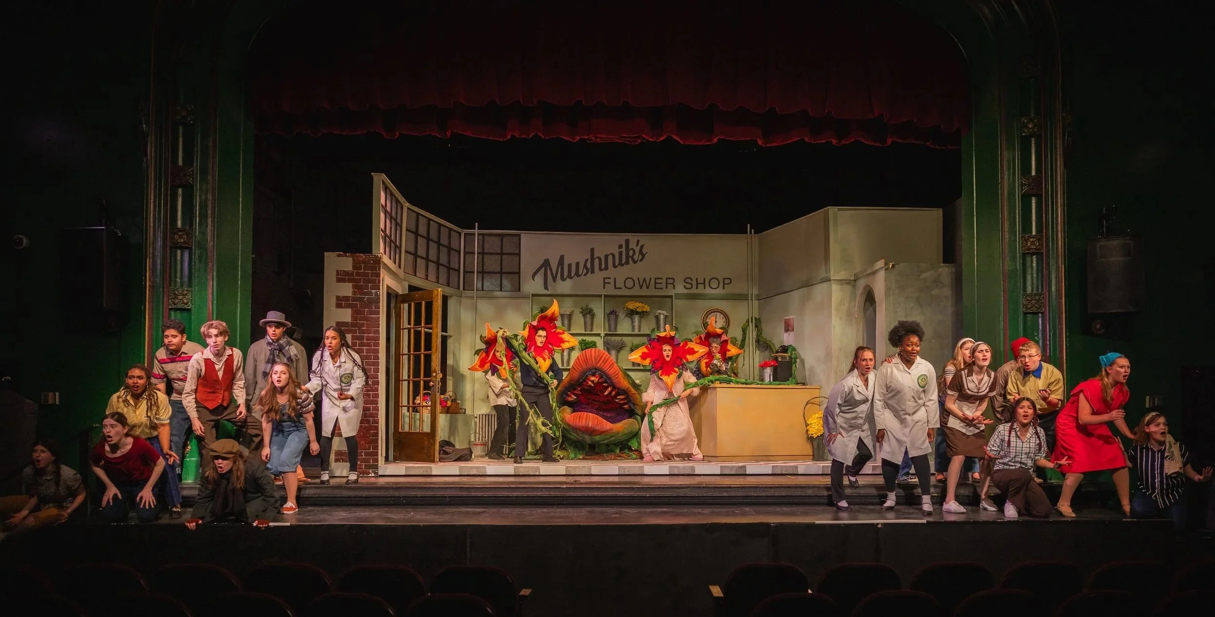 Theater stage with a cast of students performing from a play set in a flower shop, with one actor dressed as a flower and others in various costumes, in front of a backdrop with the sign 'Mushnik's Flower Shop'.