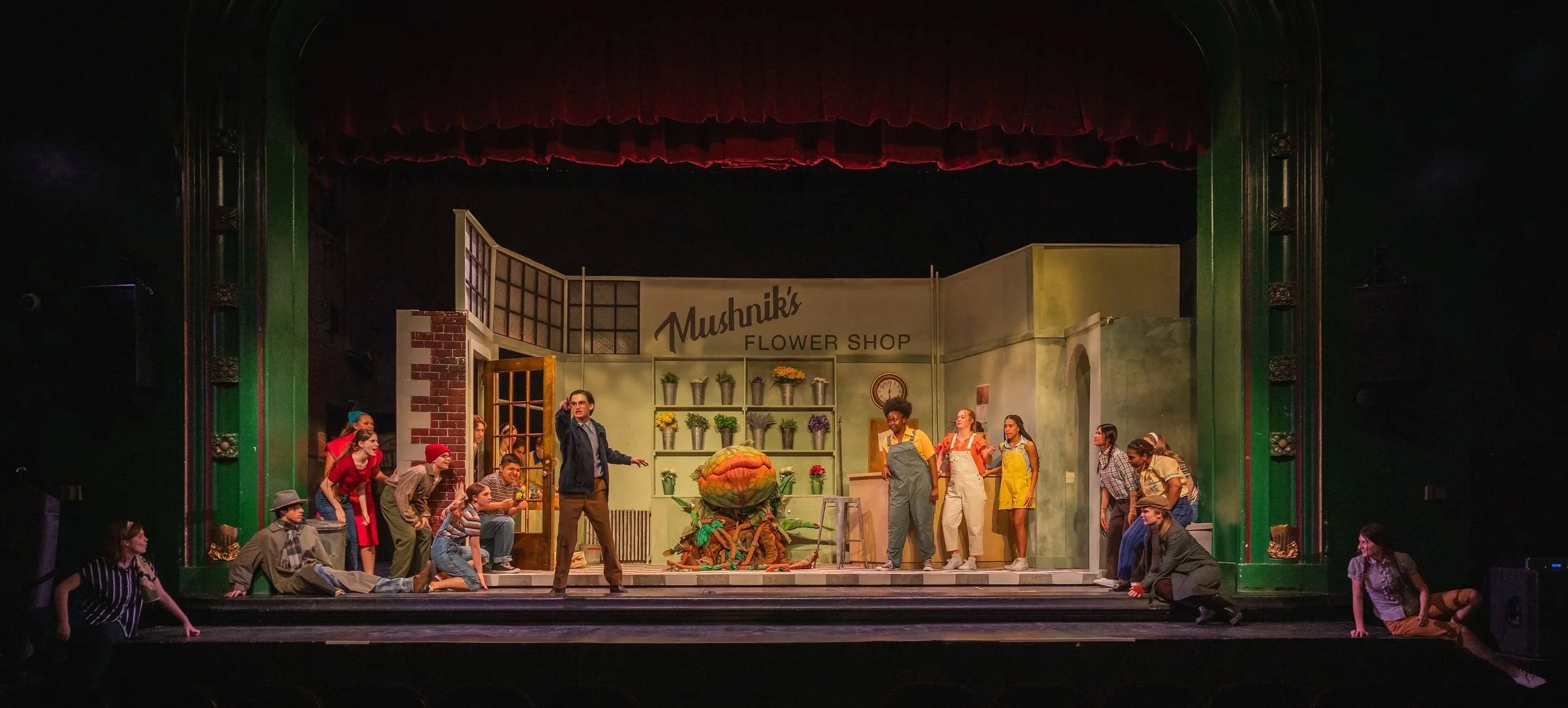 Stage scene from a theatrical production of 'Little Shop of Horros' set in 'Mushnik's Flower Shop' with actors in costume, set decorated with flowers and props, and a person speaking at center.