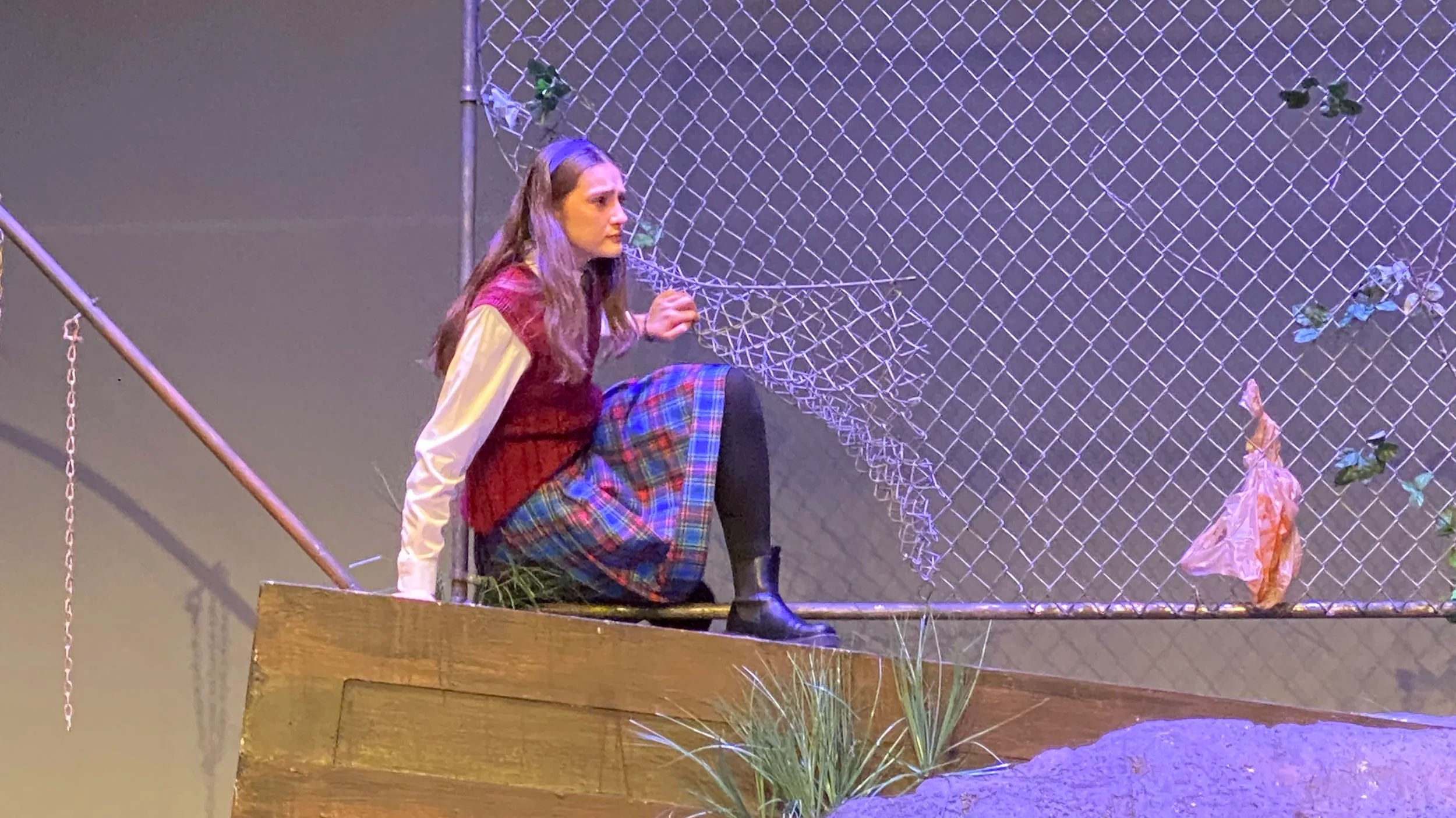 A stage scene with a woman sitting on a wooden platform in front of a chain-link fence backdrop. The fence has a sign that says 'Keep Out' and some artificial plants attached to it. The scene is part of a theatrical performance.